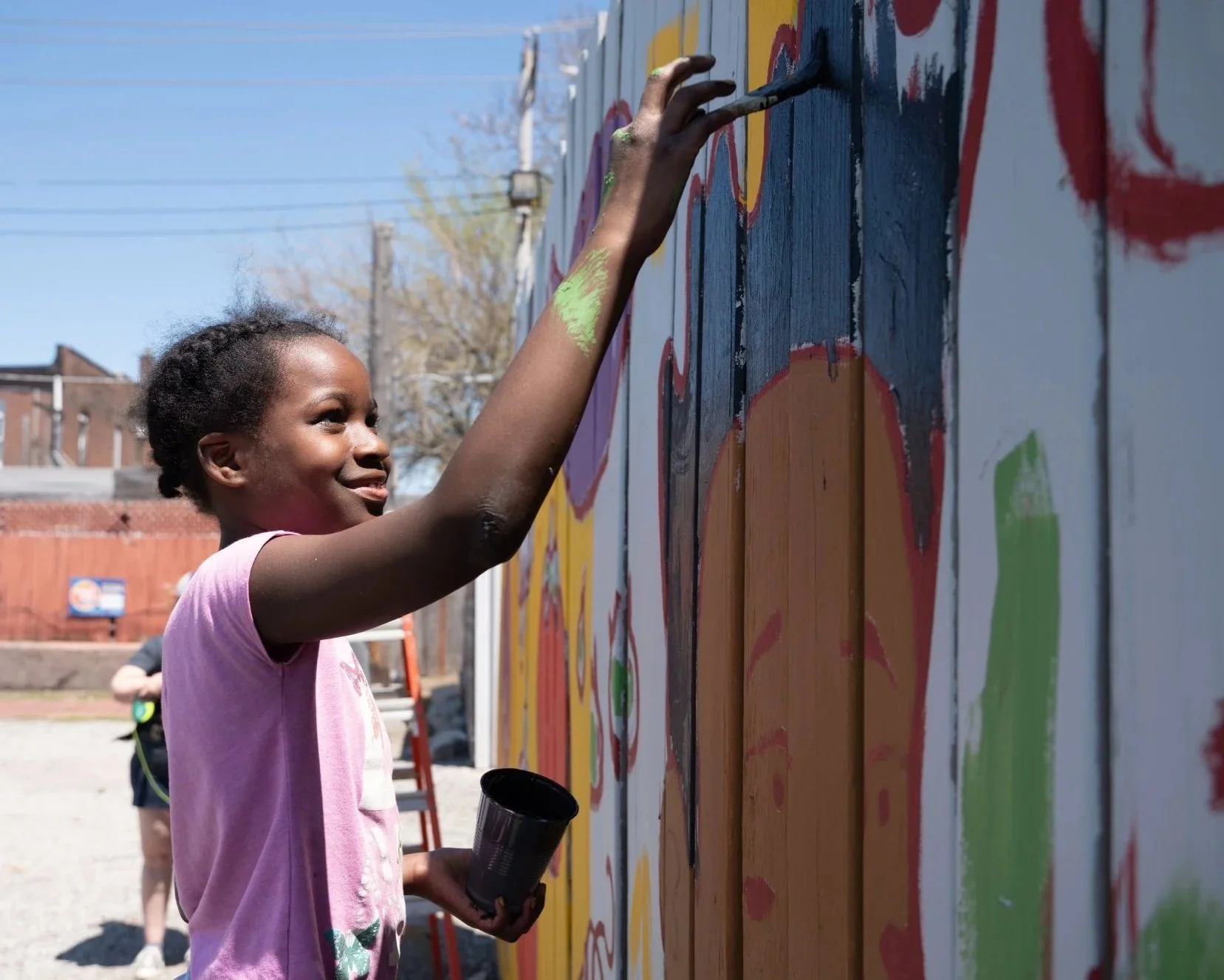 Young girl painting a colorful mural on a wooden wall outdoors, smiling as she applies paint with a brush and holding a black cup, with other children in the background.