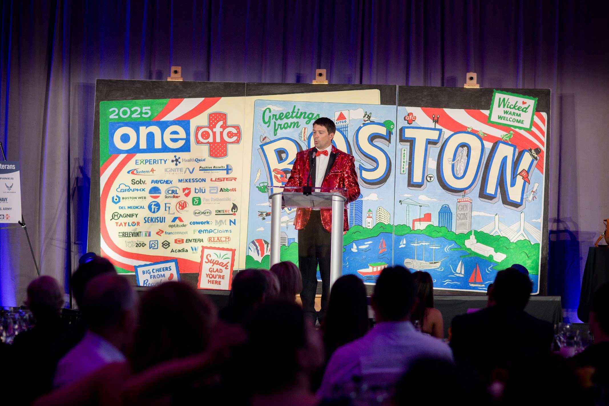 Person stands at a lectern on stage in front of a hand-painted color-by-numbers mural featuring sponsors at a boston corporate event with attendees seated at the closing night gala