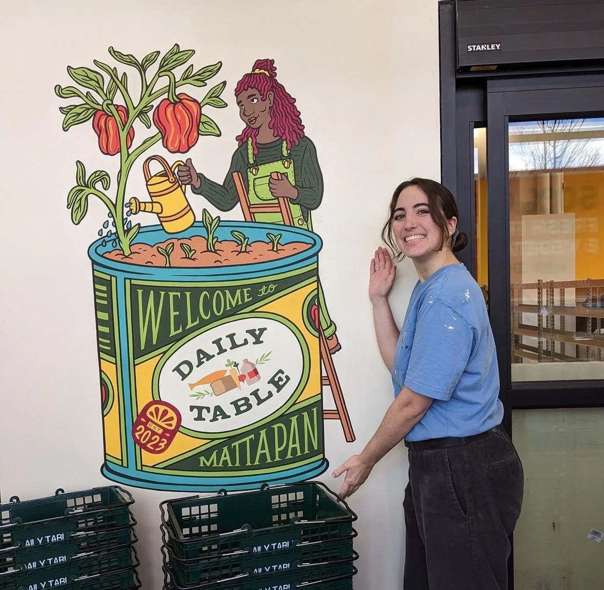 A woman smiling and posing next to a colorful mural of a woman watering a large plant in a can labeled "Welcome to Daily Table Mattapan."