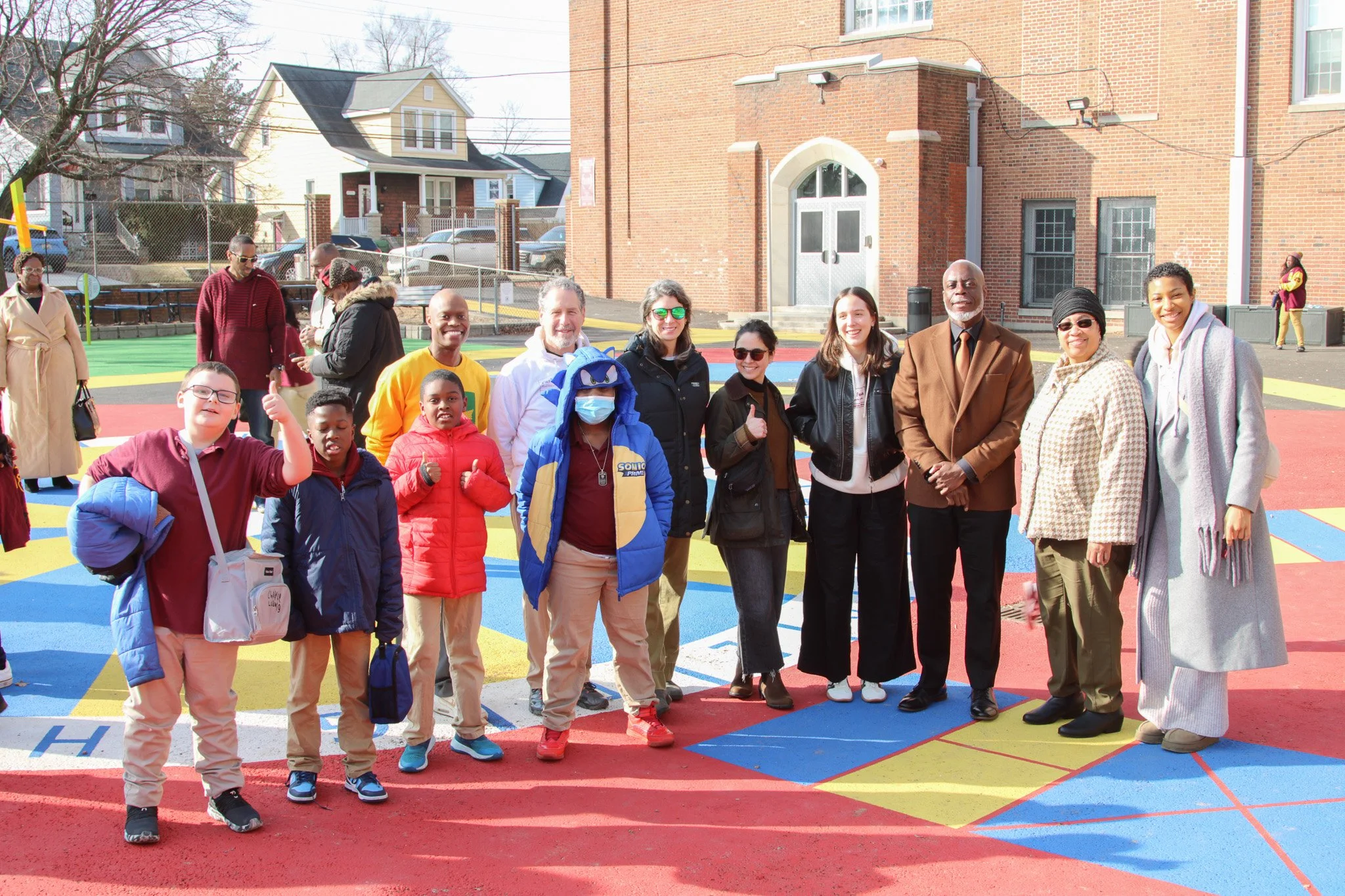 Parks & People staff and elementary and middle school students stand together on a bright, multicolored schoolyard during a ribbon-cutting celebration outside a brick school building in Northeast Baltimore.