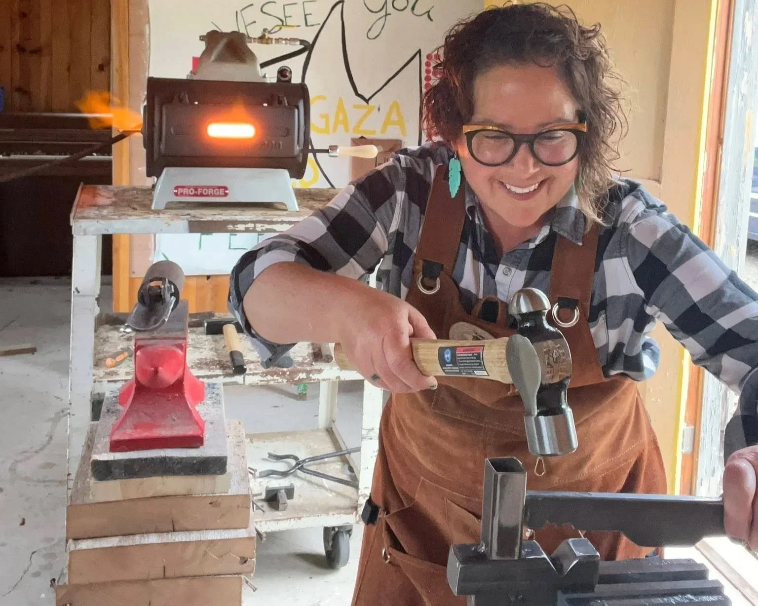 A woman smiling while hammering a piece of metal at a blacksmithing or woodworking workspace. She wears glasses, a checked shirt, and a brown apron. Behind her, there is a workbench with tools, a small forge with a glowing flame, and a wall with a mural or artwork.