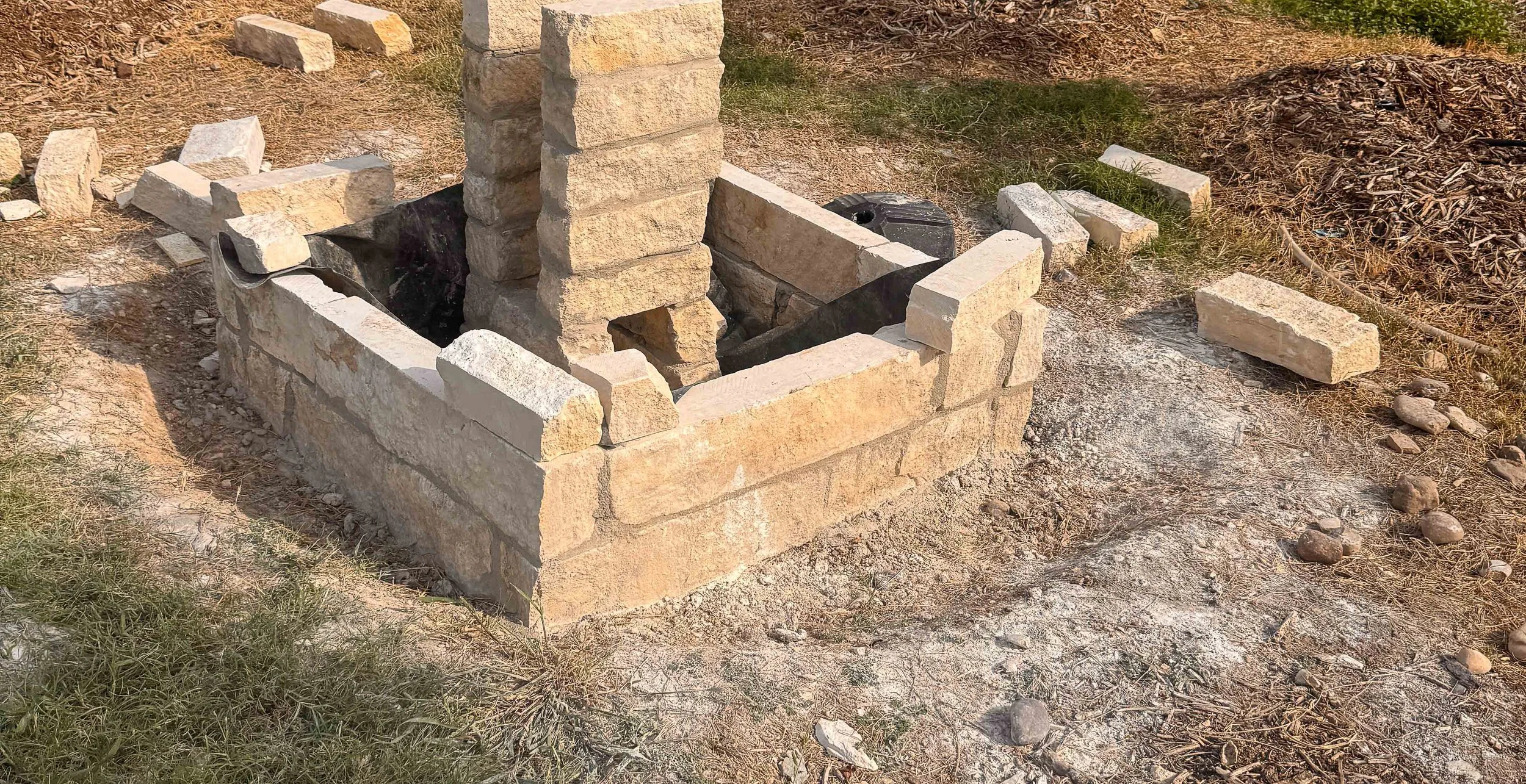 A brick fire pit under construction with some bricks stacked inside and around it, situated on dirt with patches of grass and small rocks nearby.
