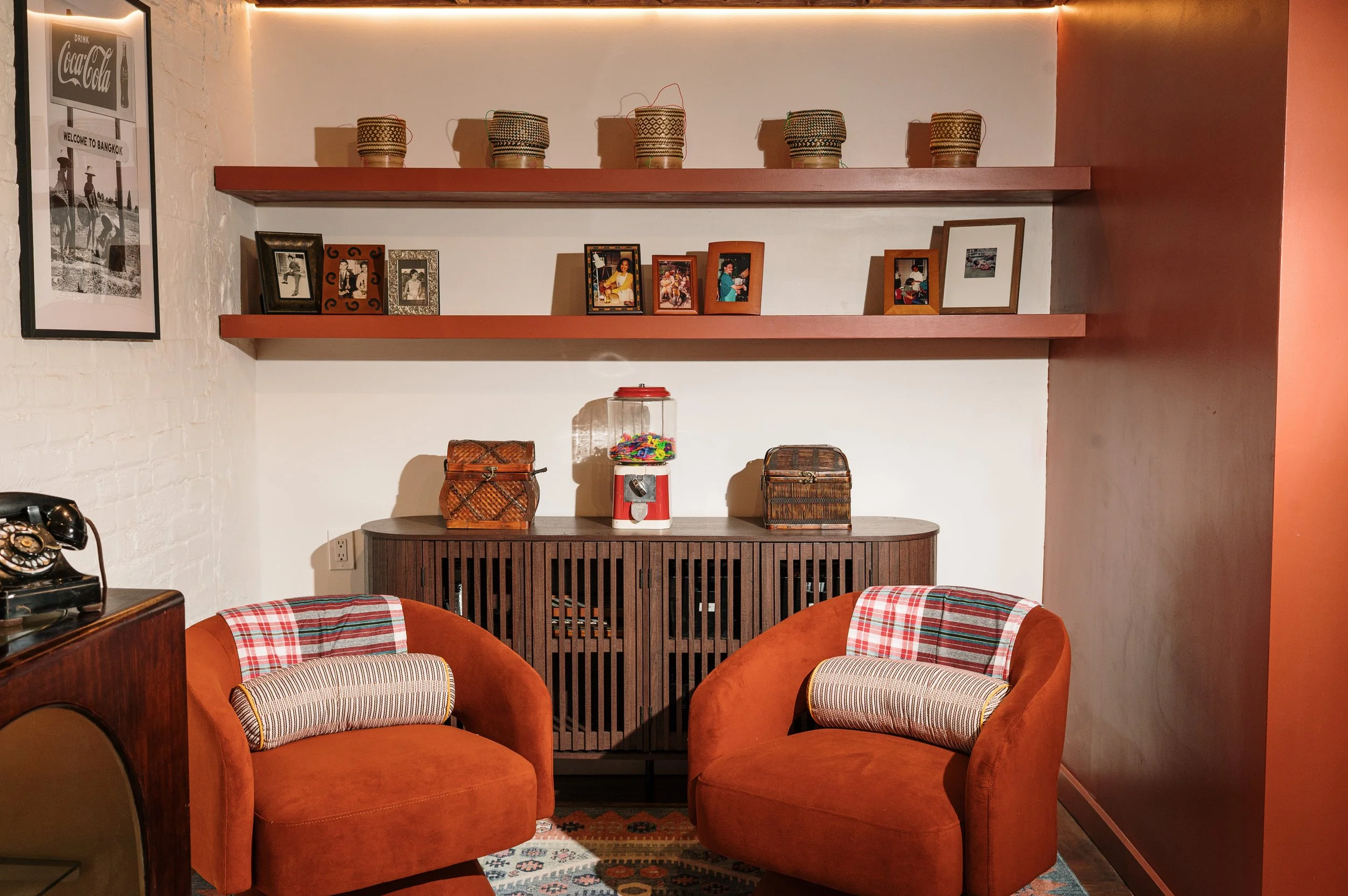 Cozy living room corner with two burnt orange armchairs, decorative pillows, and a patterned rug. Behind them, a wooden sideboard displays baskets and a gumball machine. Two pinkish-red shelves above hold framed photos and decorative jars, with a vintage black rotary phone on a side table to the left.