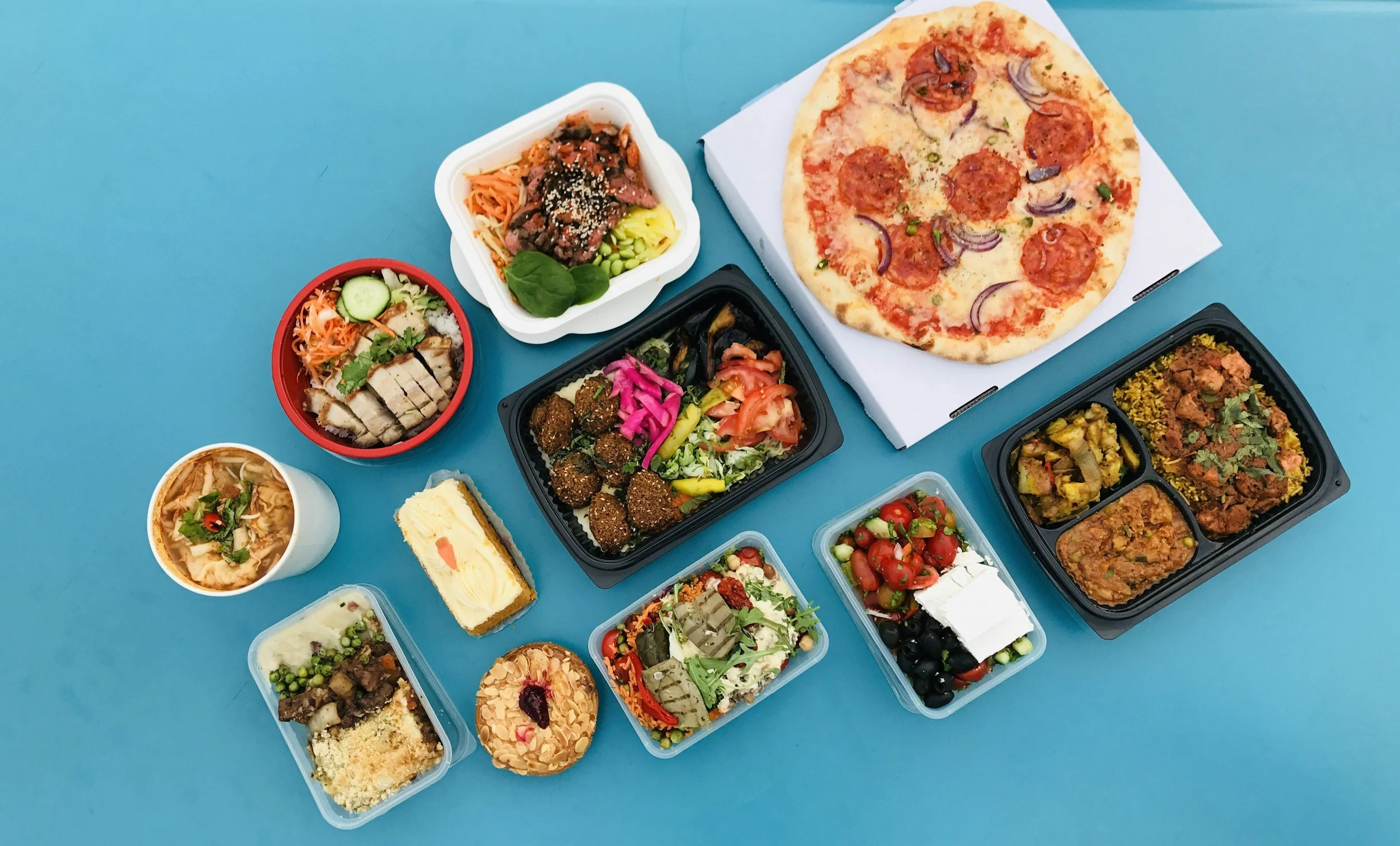 An assortment of food including a pizza with pepperoni and onions, various salads, curries, a cake, a muffin, and a cup of soup on a blue background.