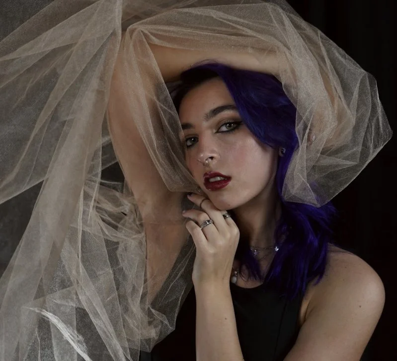 A woman with blue hair, pierced nose, and dark makeup posing with a sheer fabric draped over her head and arm, against a dark background.