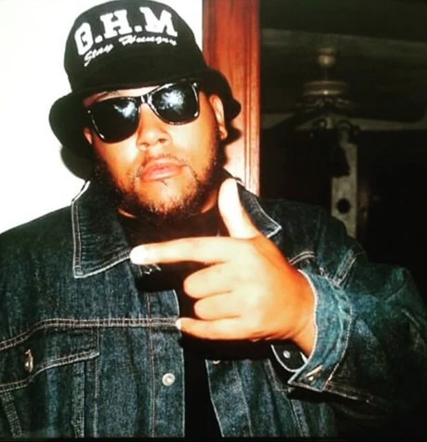 A man wearing a black cap with white text, sunglasses, and a denim jacket making a gun gesture with his hand.