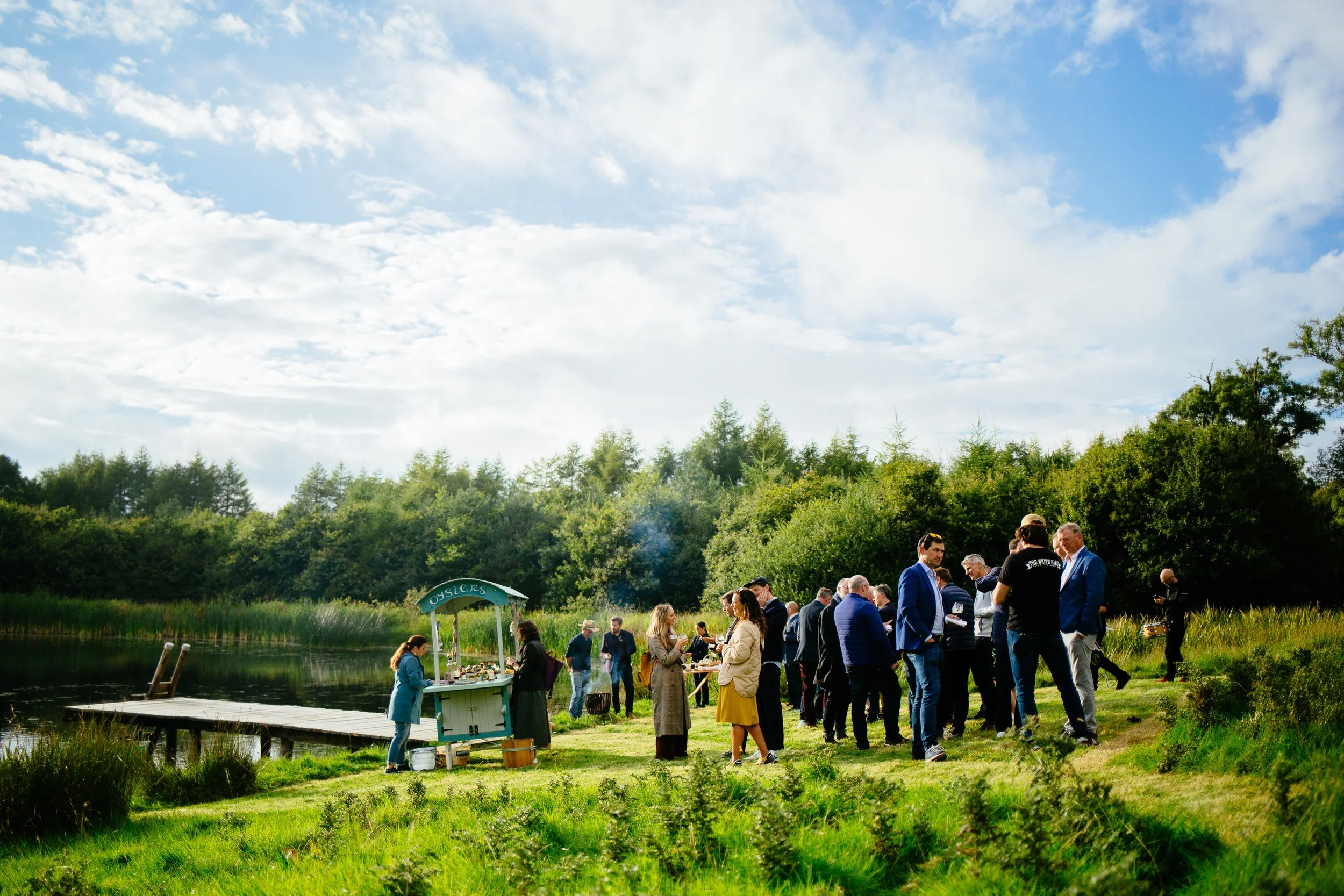 People gathering outdoors near a pond with trees and a blue sky with clouds in the background, some people are near a small table or stand by the water.