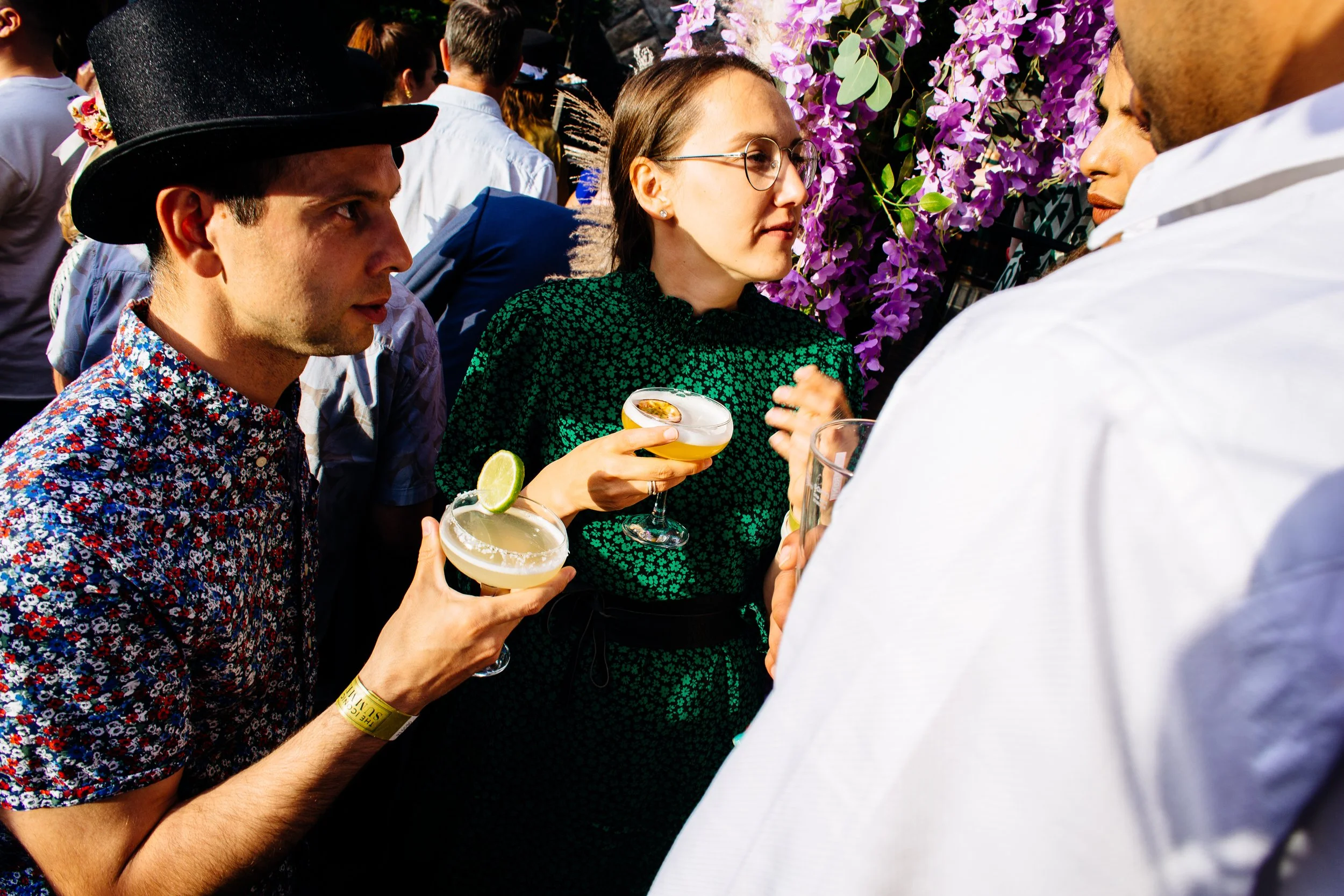 People holding cocktails at an outdoor event, with a woman in glasses and a green dress in the center, surrounded by others and purple flowering plants in the background.