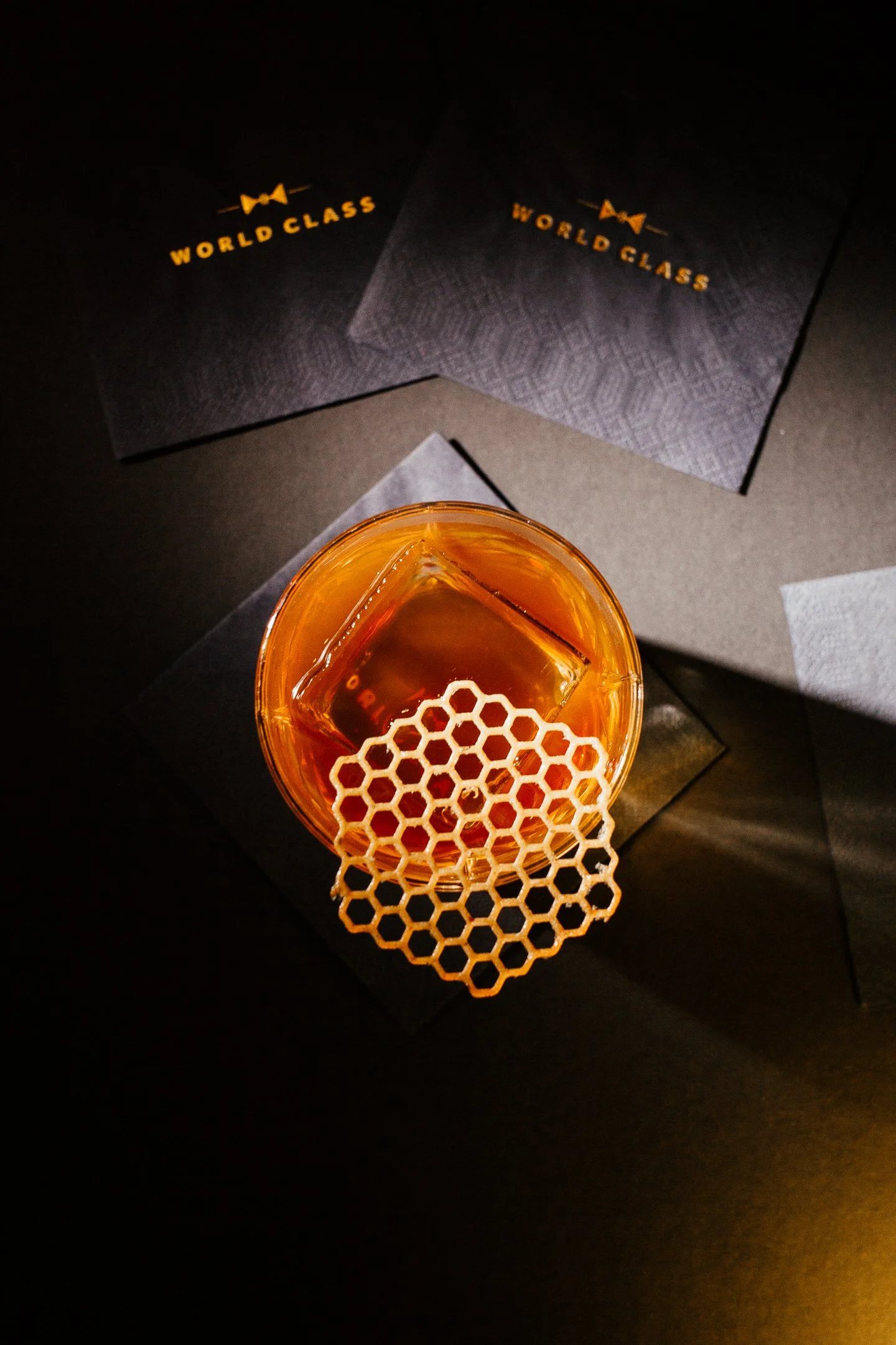 A glass of amber liquid with ice, placed on a brown surface. Two dark cards with gold text reading 'WORLDCASS' are in the background.