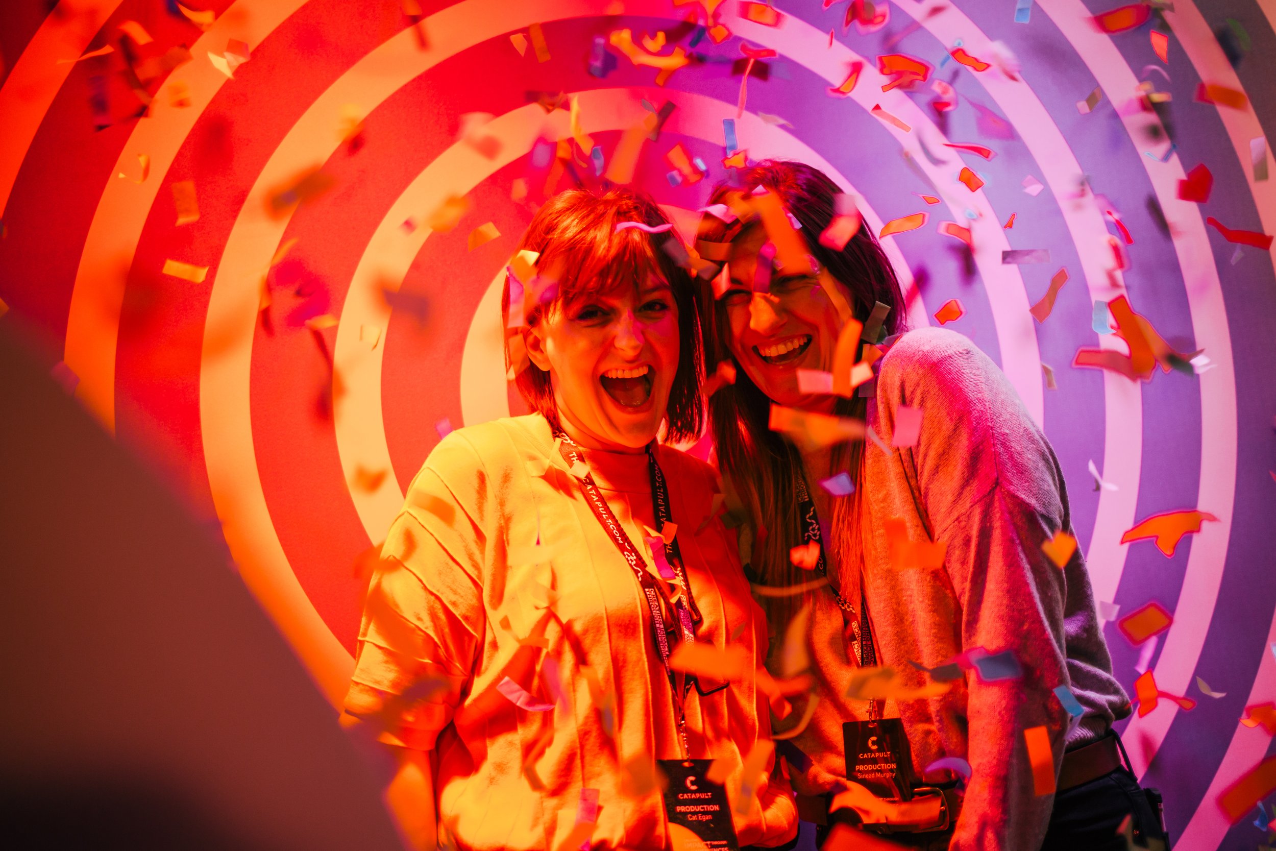 Two women celebrating with confetti, standing in front of a colorful, spiral-patterned background.