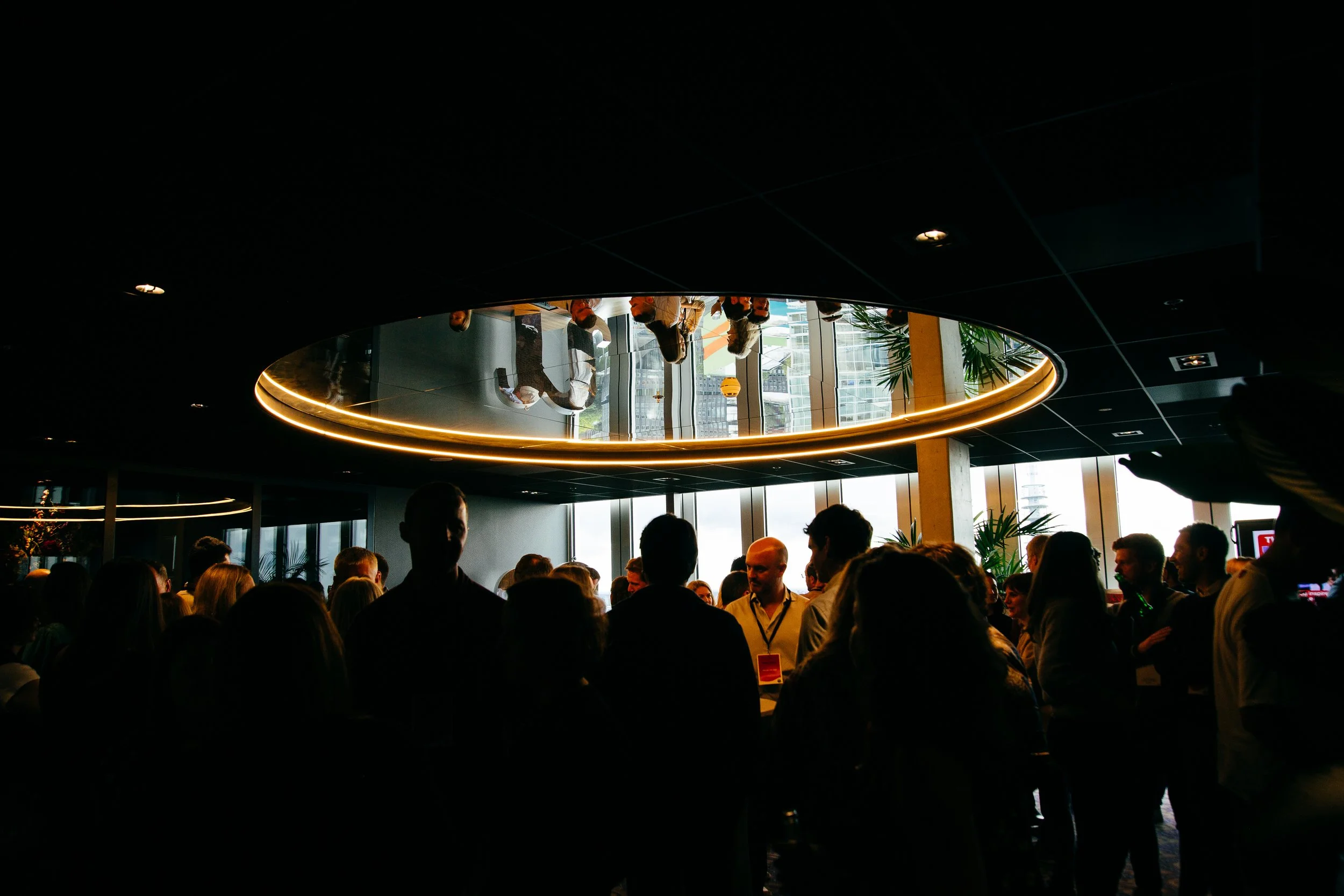 People gathered in a dimly lit indoor space with large windows, reflection in a circular ceiling mirror, modern decor, and city buildings visible outside.