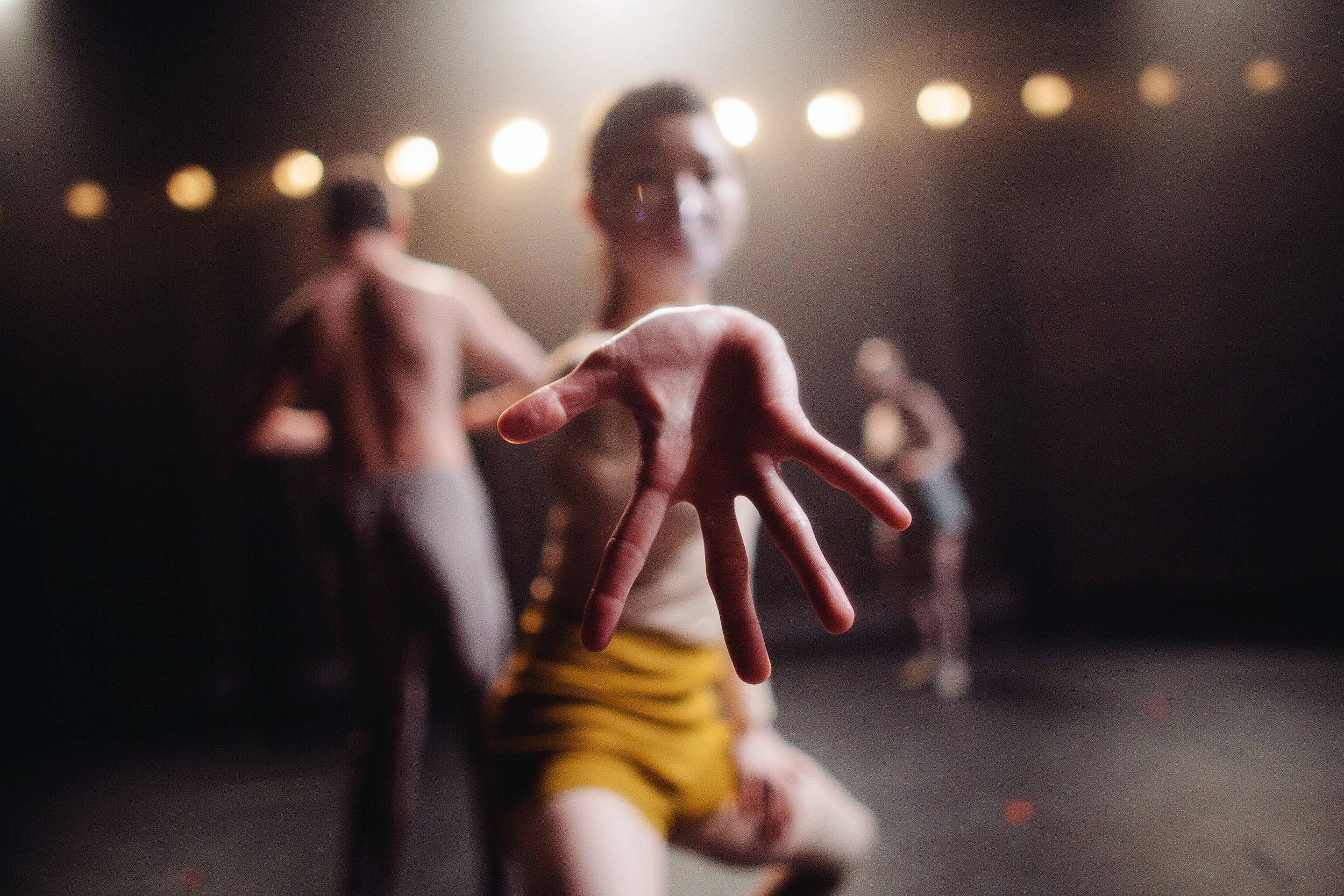 A group of dancers performing on stage with their hands extended, wearing casual attire, with a blurred background and stage lights above.