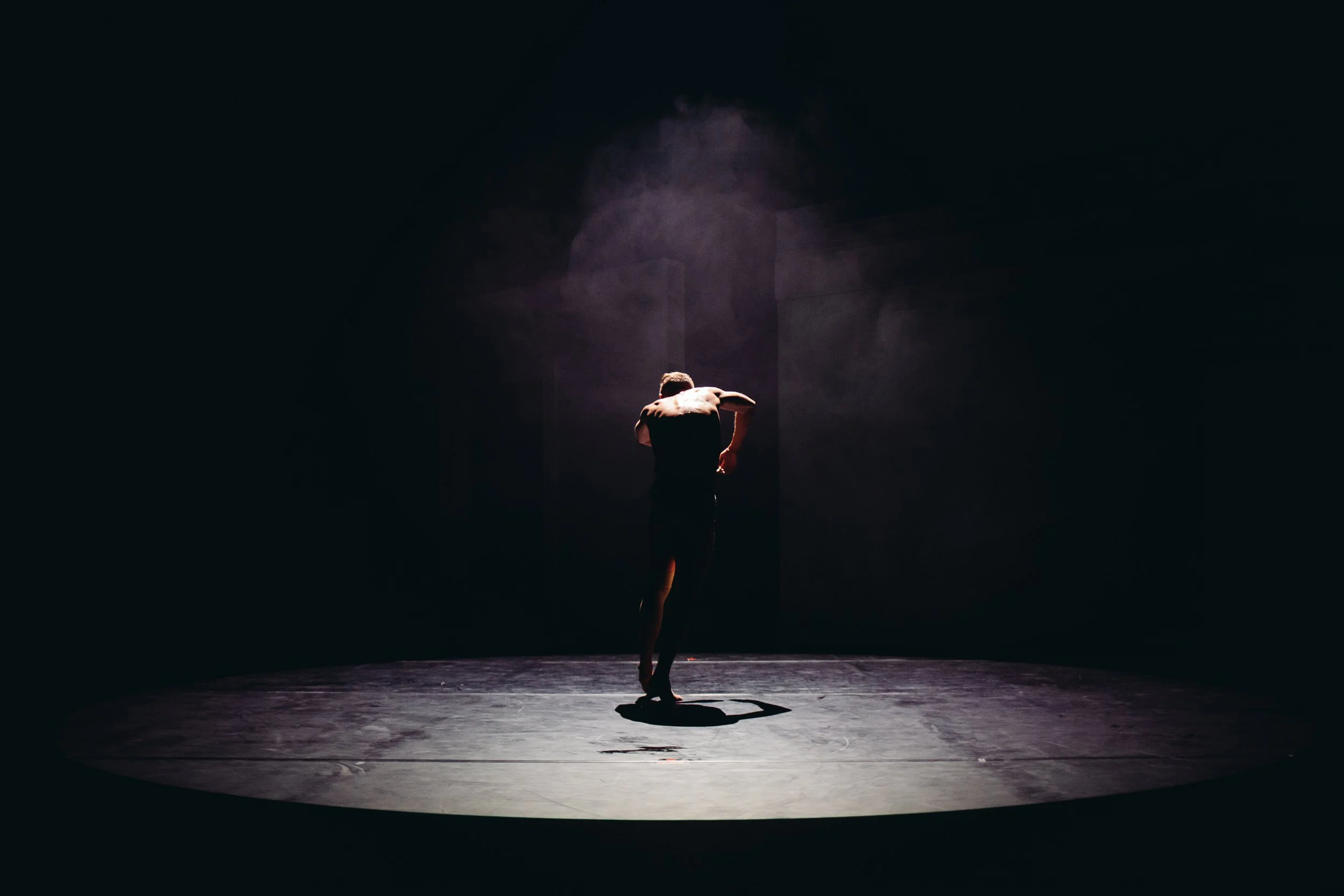 A dancer performing on a dimly lit stage, illuminated by a spotlight, with a dark background.