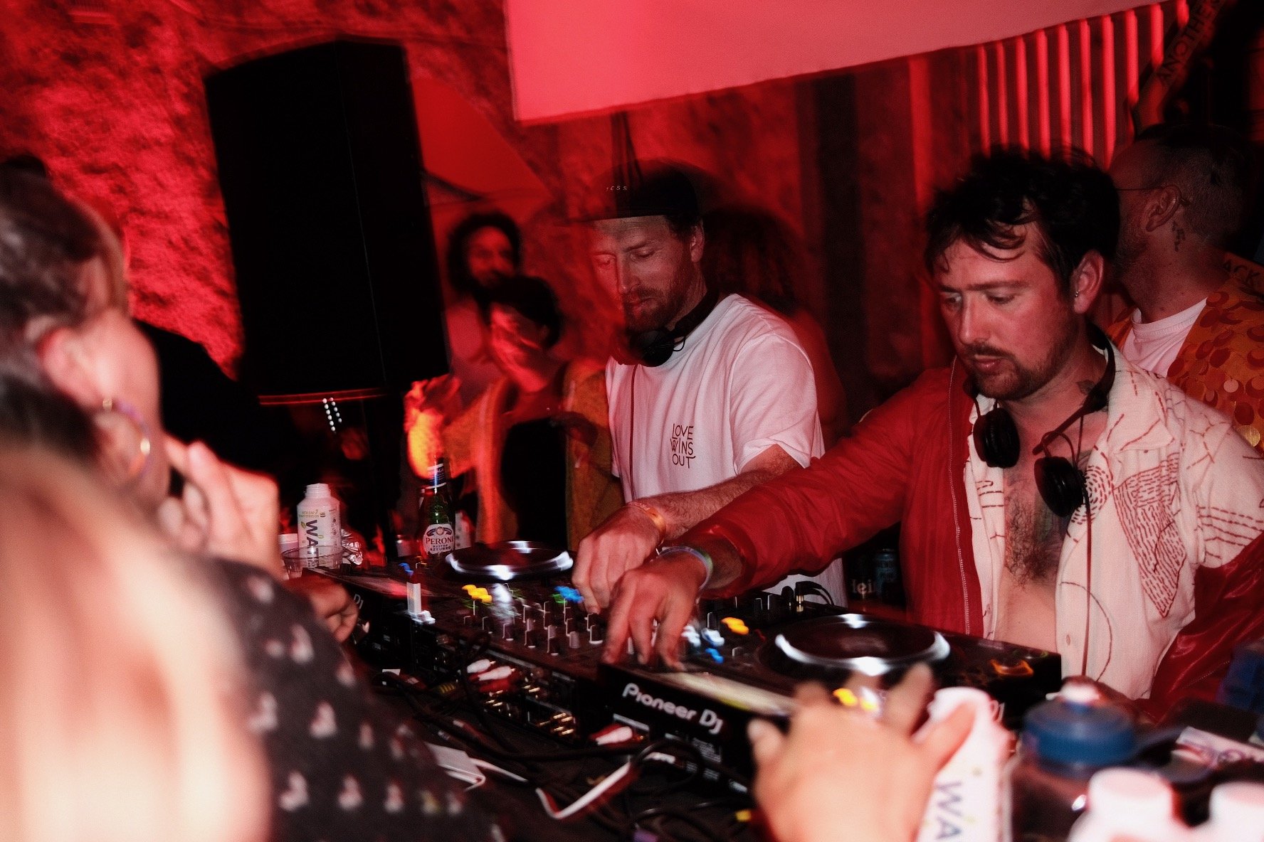 Two male DJs perform in a nightclub with reddish lighting, surrounded by a crowd. One DJ wears a white T-shirt with black headphones around his neck, and the other wears a red jacket with black headphones. Both are using DJ equipment, with people dan