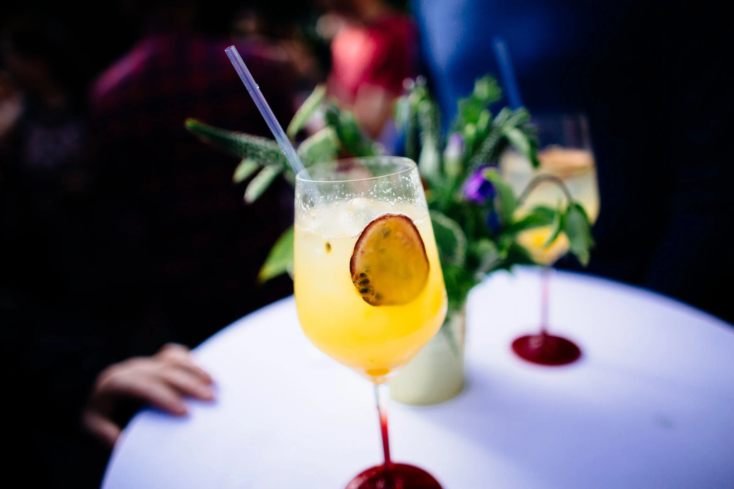 A colorful cocktail drink with a pineapple slice garnish and a blue straw, with blurred plants and a person in the background.