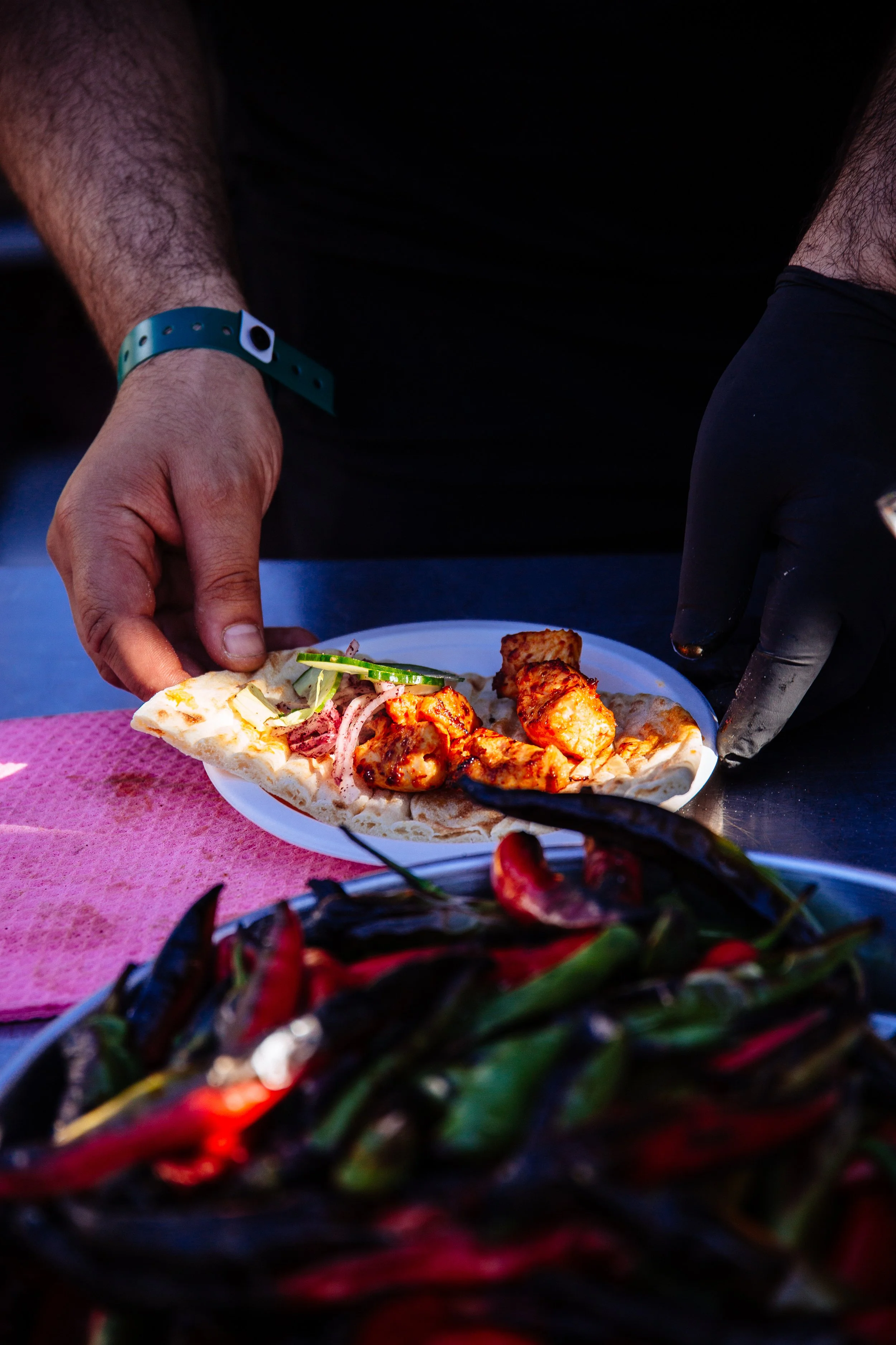 A person serving food on a white plate with a slice of pizza topped with chicken and vegetables, garnished with cucumber slices and onions at a food setting. In the foreground, there is a colorful dish with chili peppers and vegetables. The person is