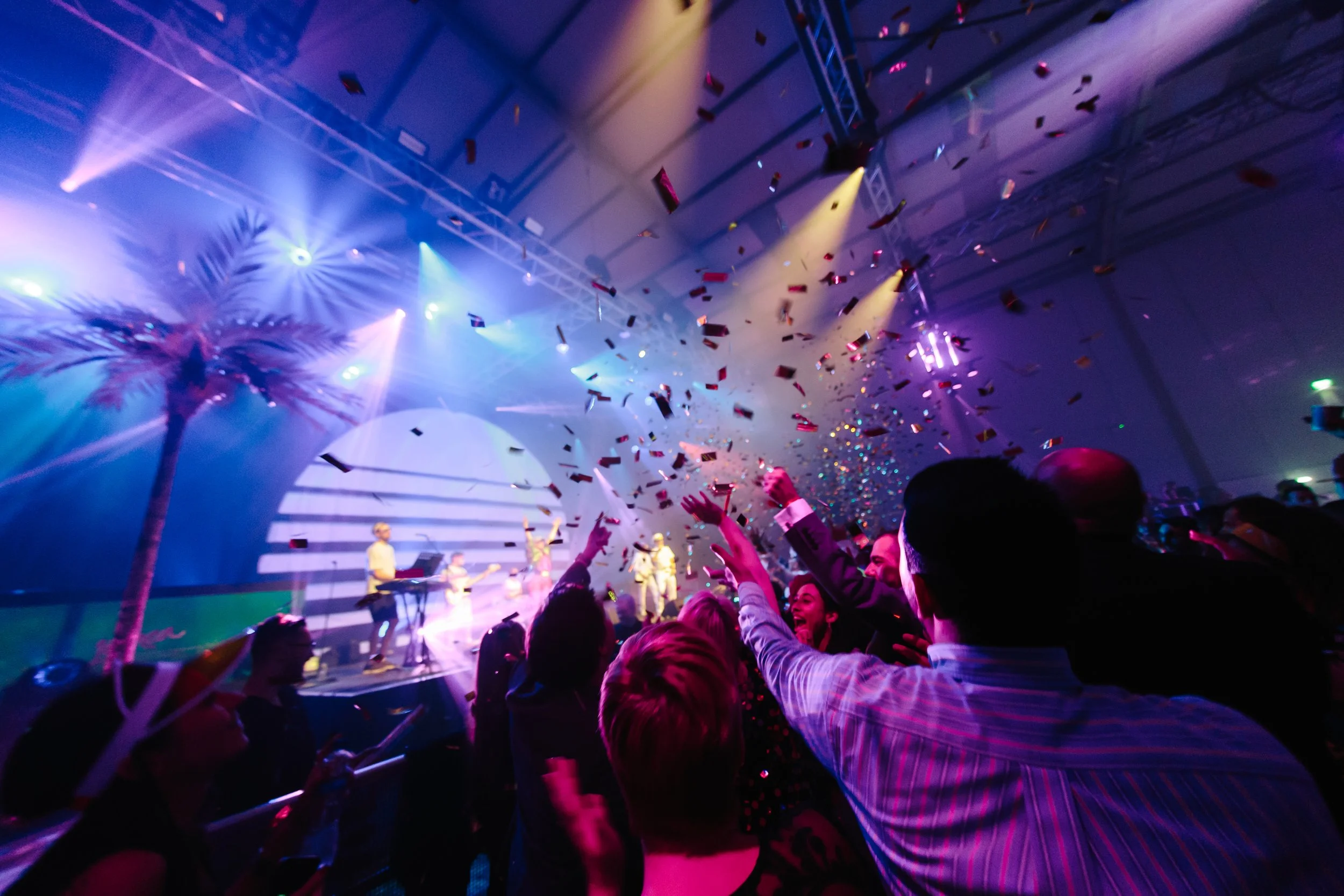 A lively indoor party with colorful lights, confetti falling, a tropical palm tree decoration, and people dancing and celebrating.
