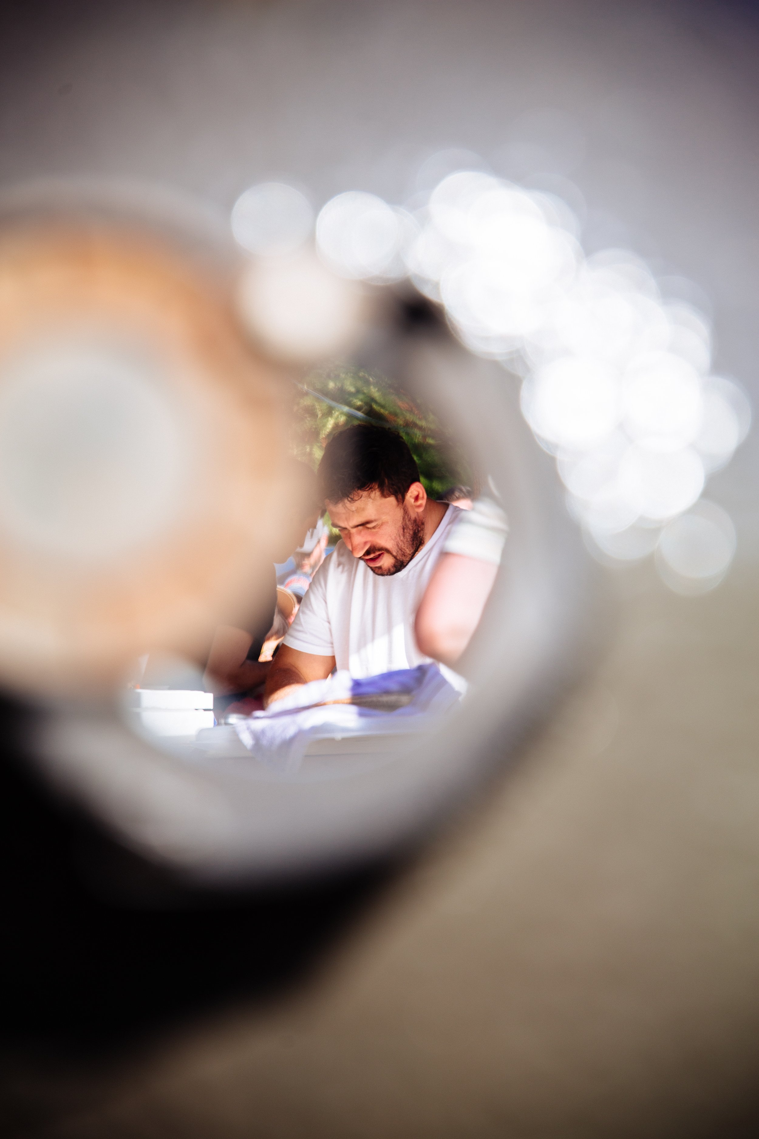 A person with dark hair and beard sitting at a table, smiling, seen through a circular hole in an object, with bokeh lights in the background.