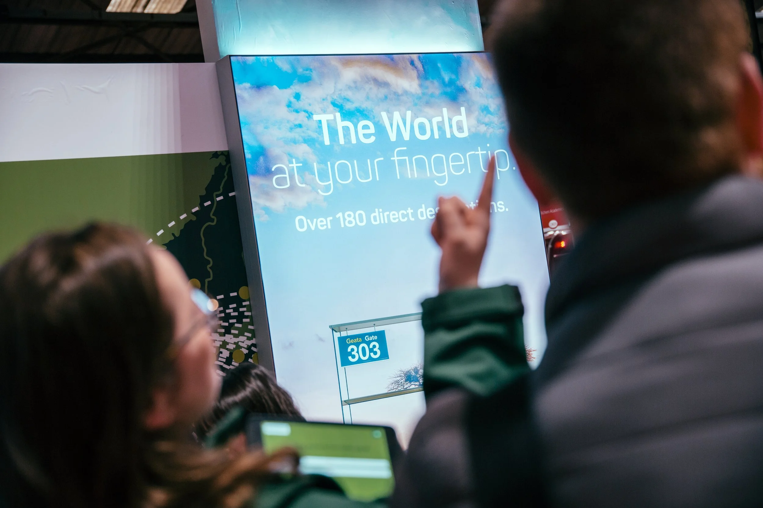 People waiting at an airport terminal with a display screen showing information about The World at your fingertips, over 180 direct destinations, and gate 303 at Geata.