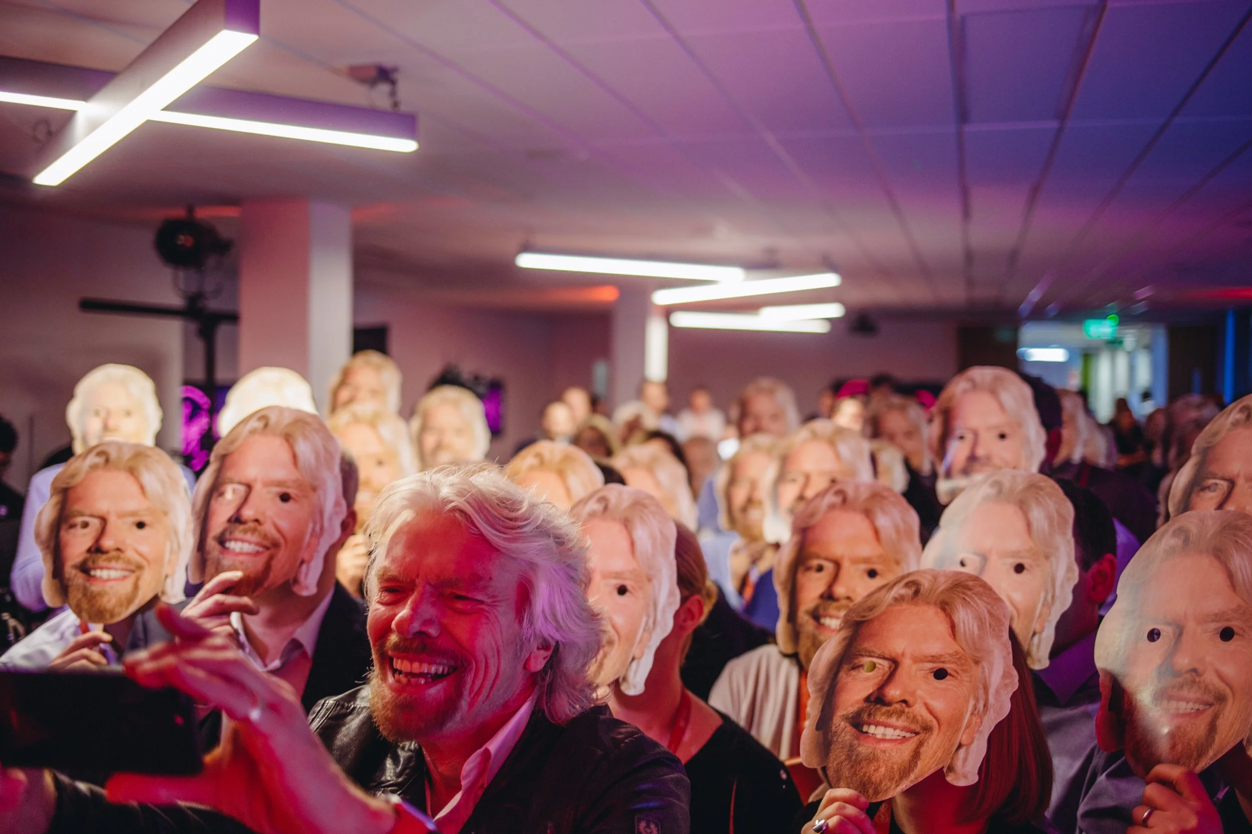 Crowd of people holding cutouts of Donald Trump’s face at an event with purple and blue lighting.
