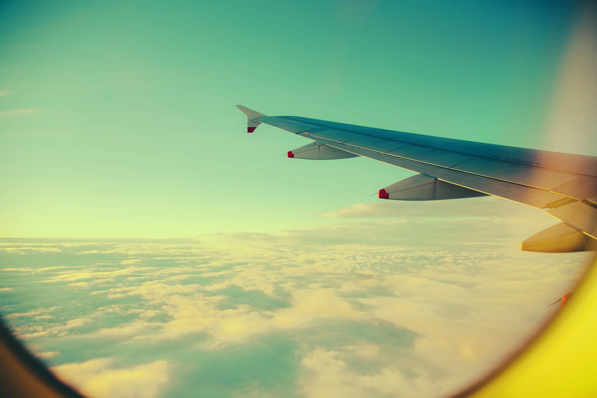 View of airplane wing flying above clouds, seen through an airplane window with a yellowish tint.