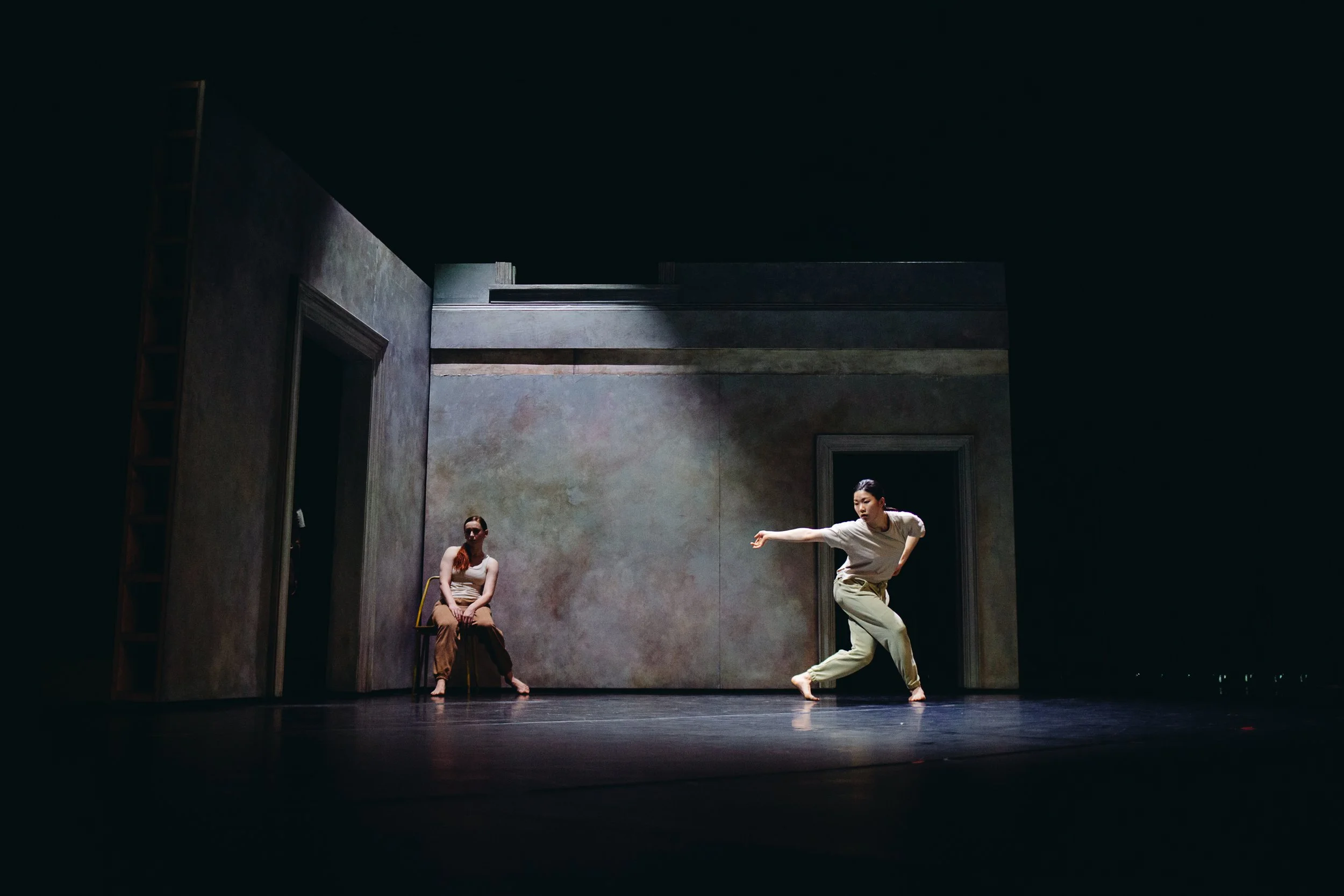 Two women performing a dance or theatrical piece on a dimly lit stage with a plain back wall. One woman is seated on a chair, and the other is dancing or posing dynamically.