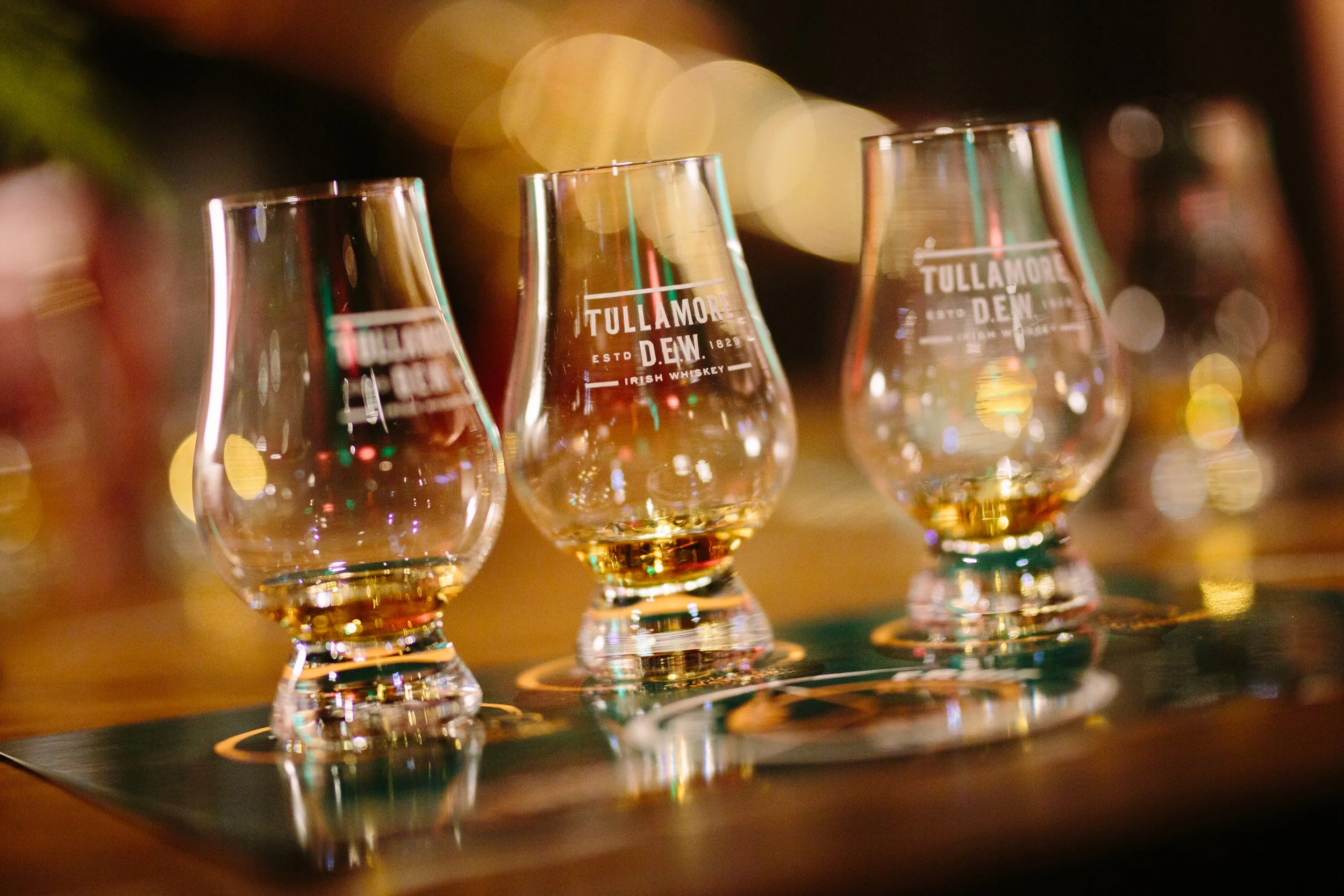 A row of three whiskey glasses labeled Tullamore D.E.W Irish Whiskey, with colorful bokeh lights in the background.