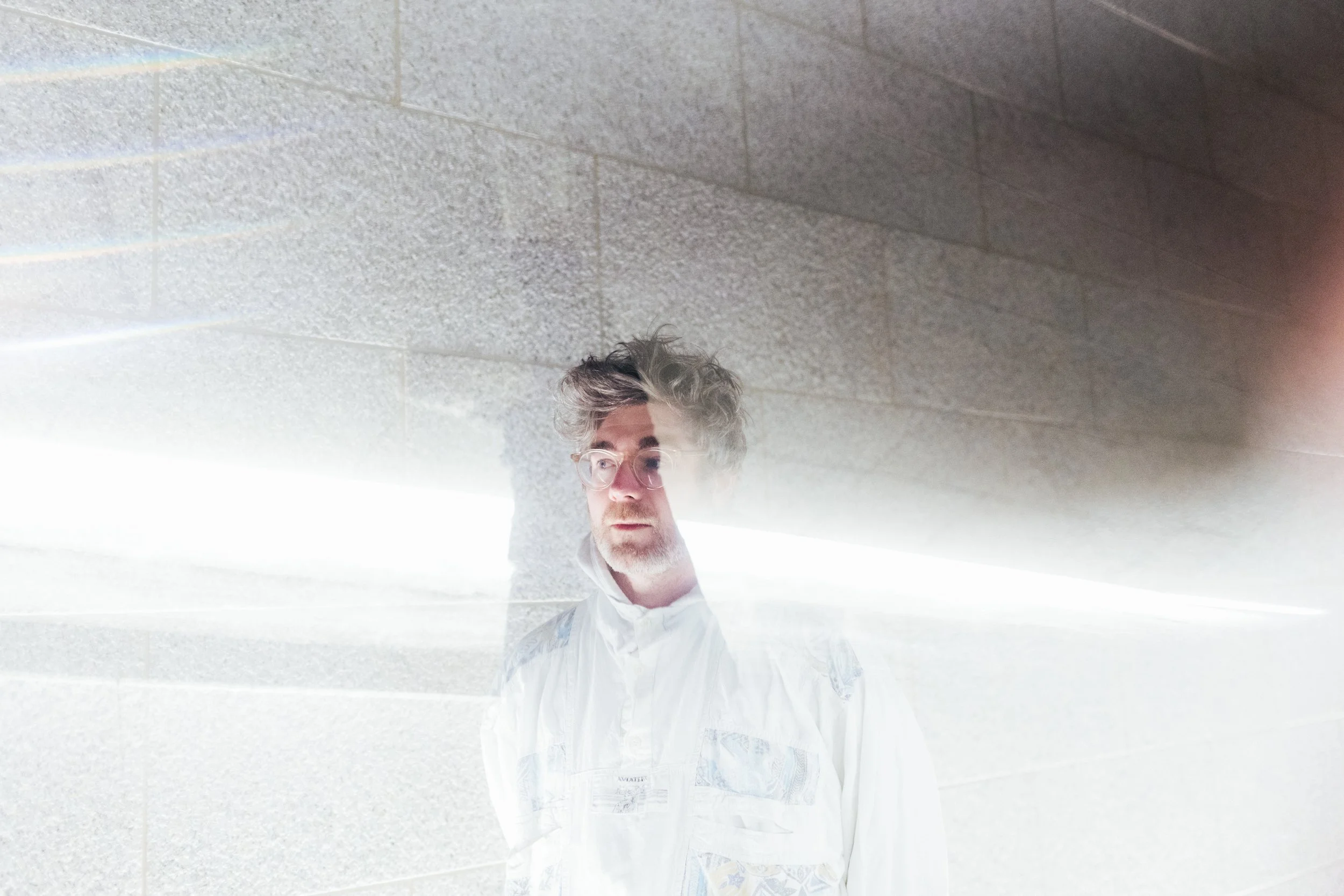 A man with glasses and curly hair looking at a reflective surface on a wall.