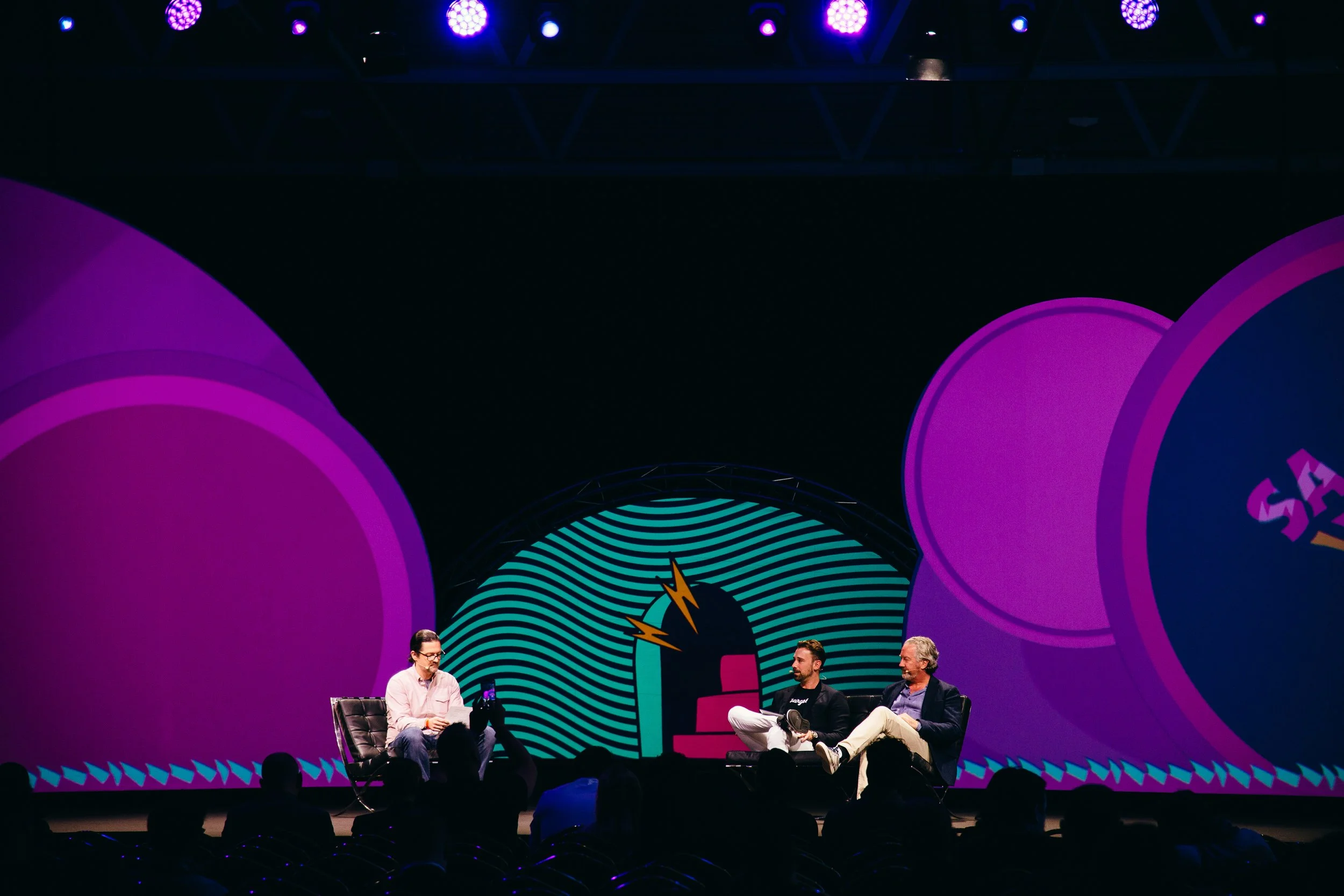 Three men sitting on chairs on stage, participating in a panel discussion, with a colorful, circular LED screen background and stage lighting.