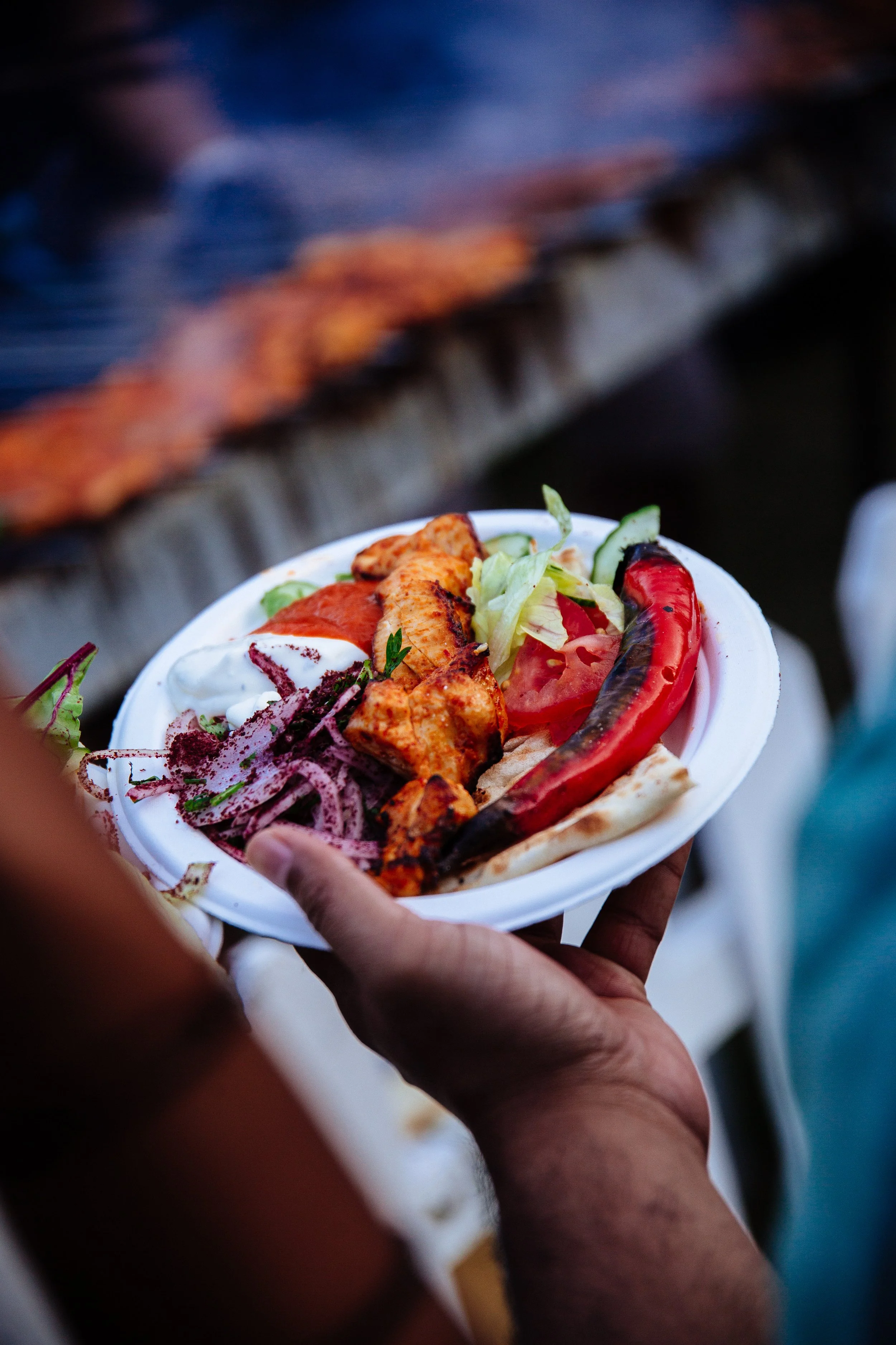 A person holding a white bowl with grilled chicken, sliced tomatoes, lettuce, grilled red chili, and purple cabbage salad, with a barbecue grill in the background.