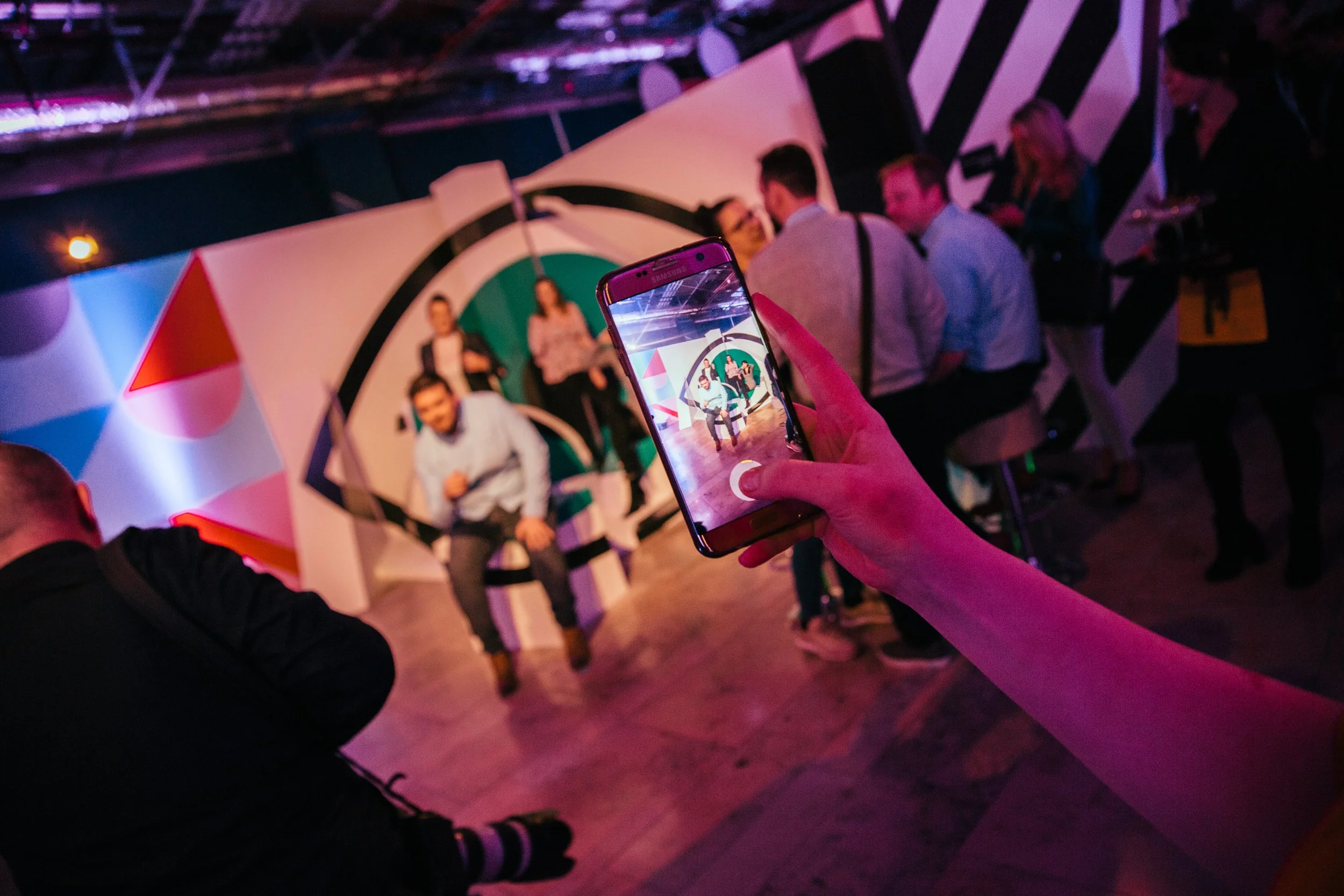 Person in foreground taking a photo of a group of people posing in front of a dartboard-style backdrop on a stage at an event or party.