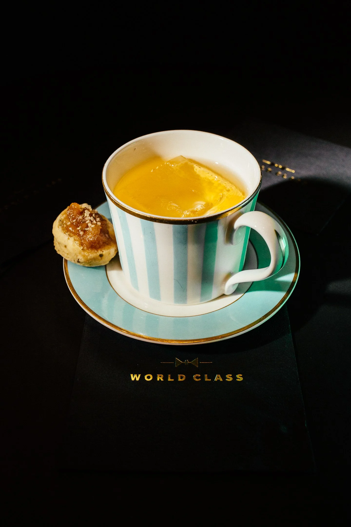 A ceramic mug with blue and white stripes containing a yellow liquid, placed on a matching saucer with a cookie next to it, on a black surface with the words "WORLD CLASS" in gold lettering.