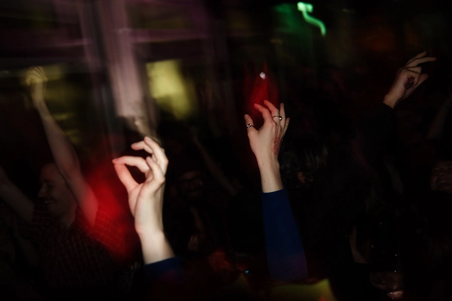 People dancing and raising their hands in a dark indoor setting, illuminated by colorful neon lights.