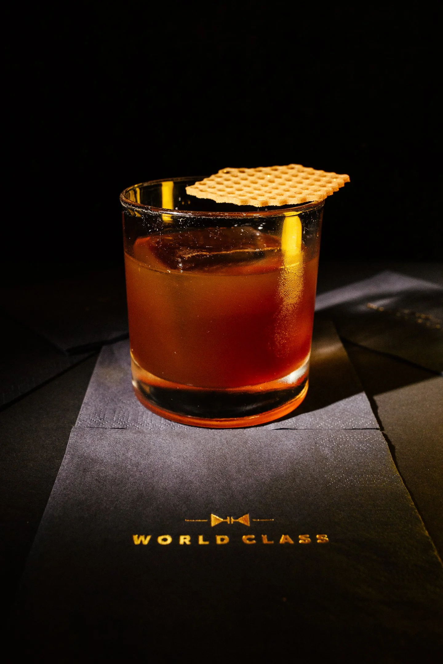 A glass of amber-colored whiskey with a piece of waffle on the rim, placed on a black World Class menu or placemat.