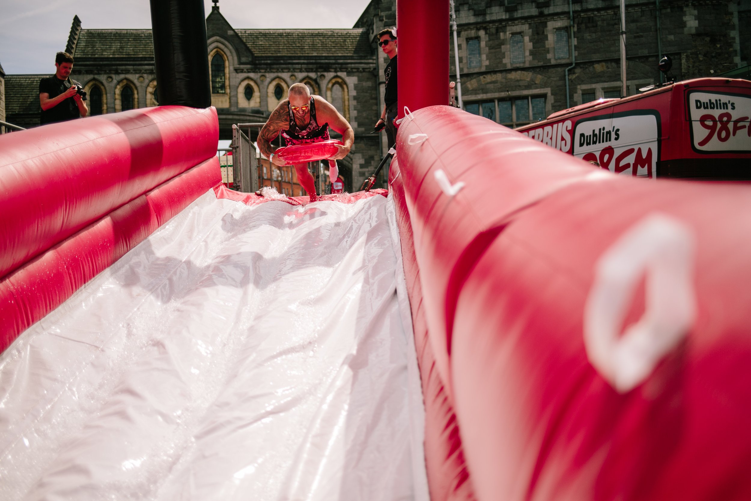A man with tattoos and sunglasses sliding down an inflatable water slide with a red exterior and white interior, while another man and a woman watch nearby during daytime in an urban area with historic buildings in the background.