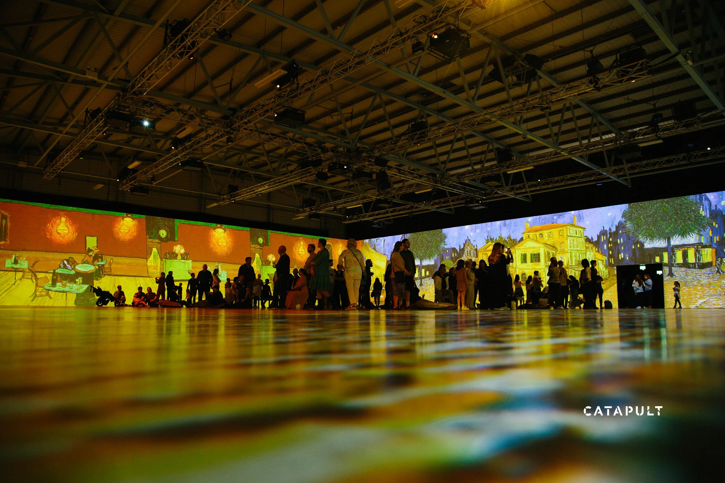 An indoor event space with a dark ceiling and exposed metal beams, filled with people standing and walking. Large digital screens display vibrant images of Vincent van Gogh's paintings, creating an immersive art experience.
