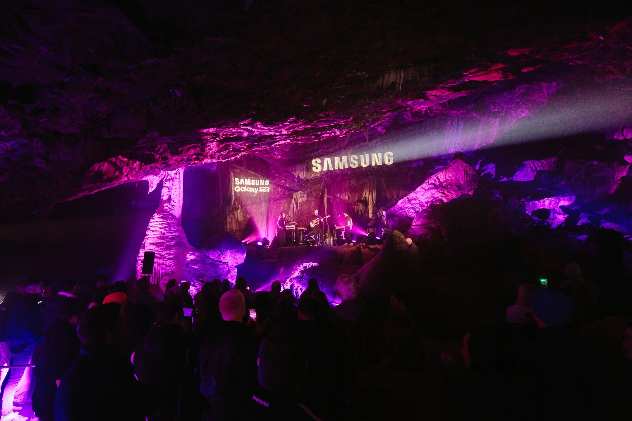 Concert inside a cave with purple and pink lighting, featuring musicians playing guitars and drums, with 'SAMSUNG' and 'Samsung Galaxy S23' logos projected on the rock wall.