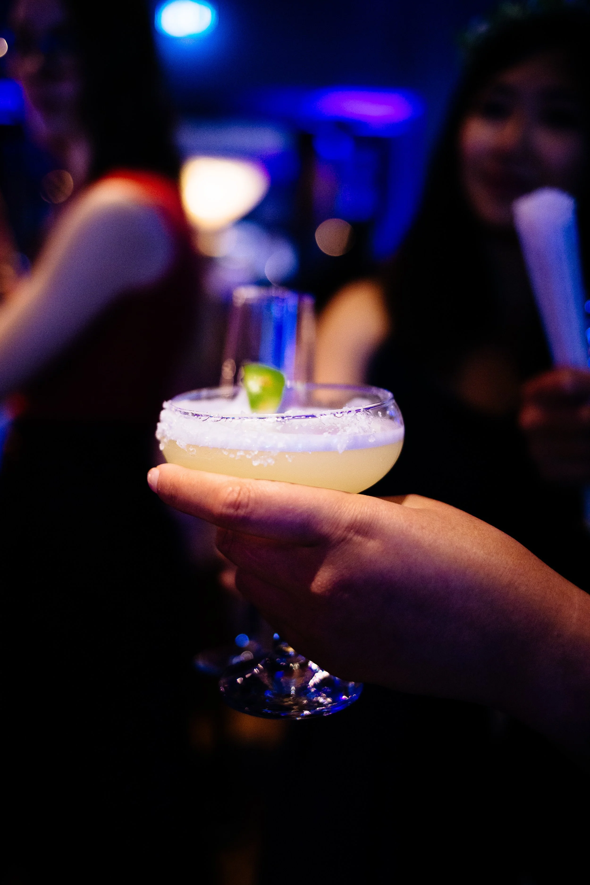 A person holding a cocktail glass with a salted rim and lime garnish in a dark, neon-lit bar or club setting, with blurred people and colorful lights in the background.