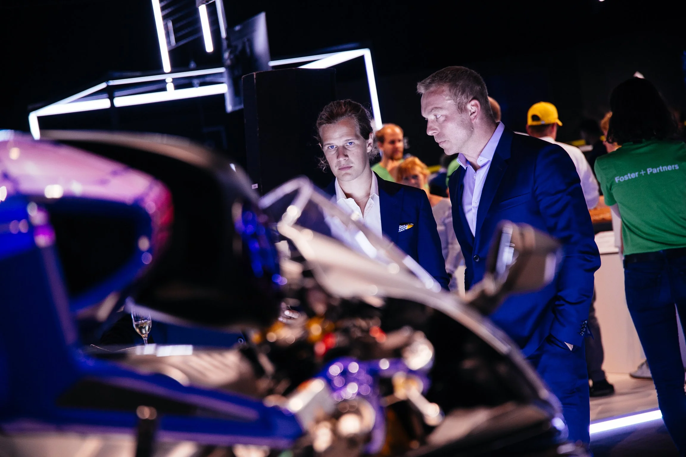 Two men in suits examining cars at an indoor exhibition, with blurred background including people and lighting.