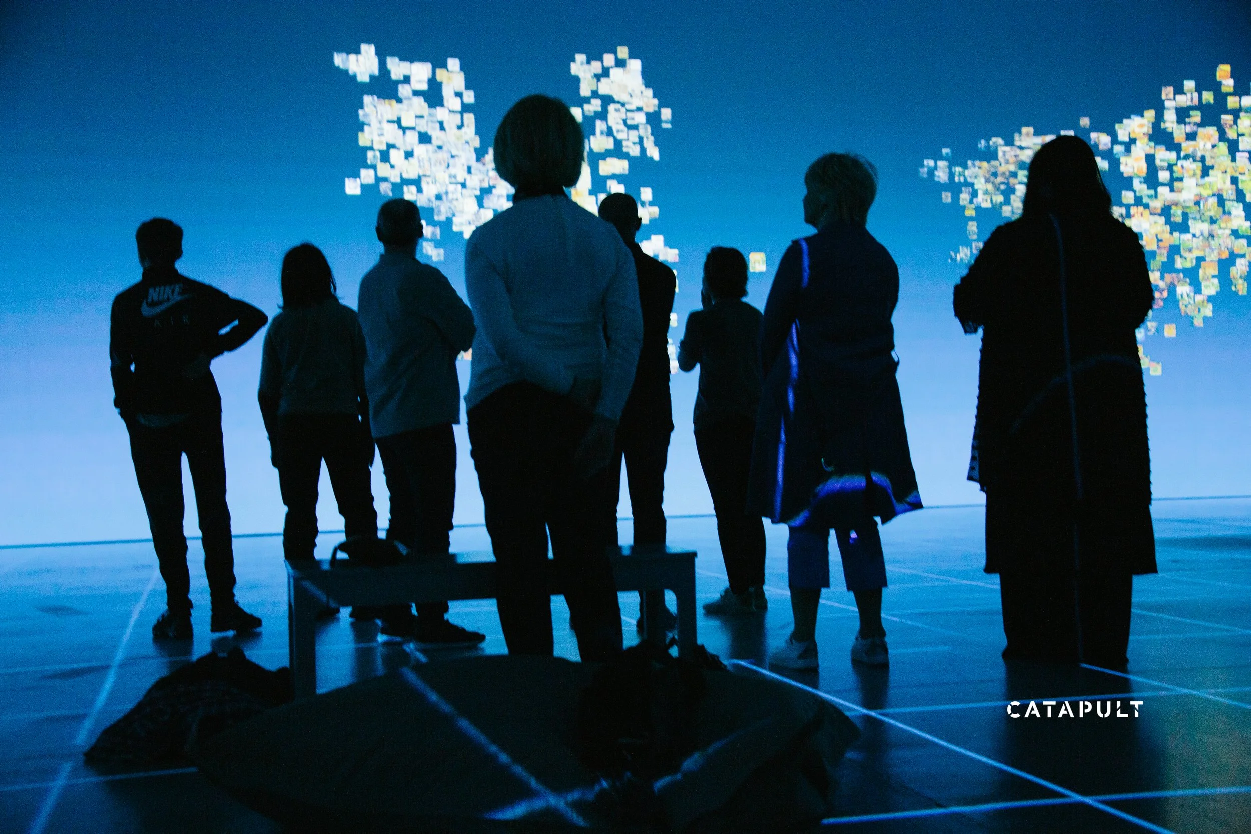 Silhouettes of people standing in a dark room watching a digital display with pixelated images and text, including the word 'CATAPULT' at the bottom right corner.