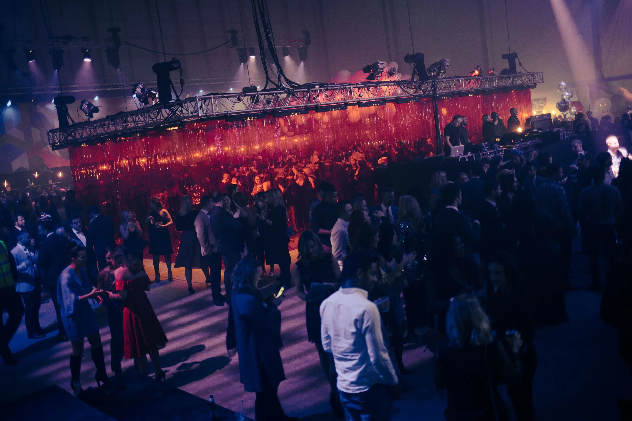 A crowded indoor event with people dancing and socializing under colorful lights, and a DJ or music setup visible on the right side.