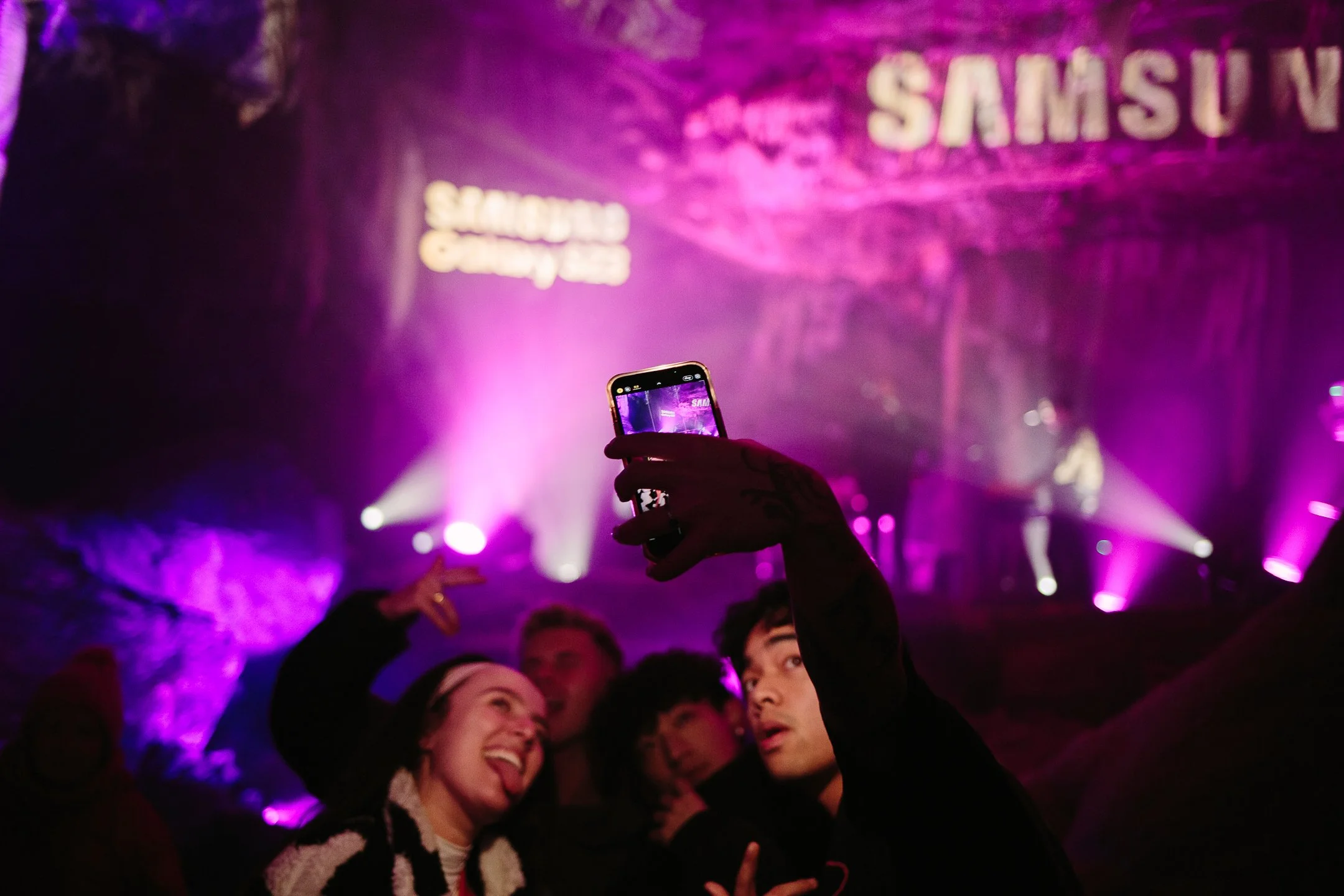A crowd at a party or concert with people taking pictures and enjoying the event, illuminated by purple and pink stage lights, with a large sign reading 'SAMSUNG' in the background.