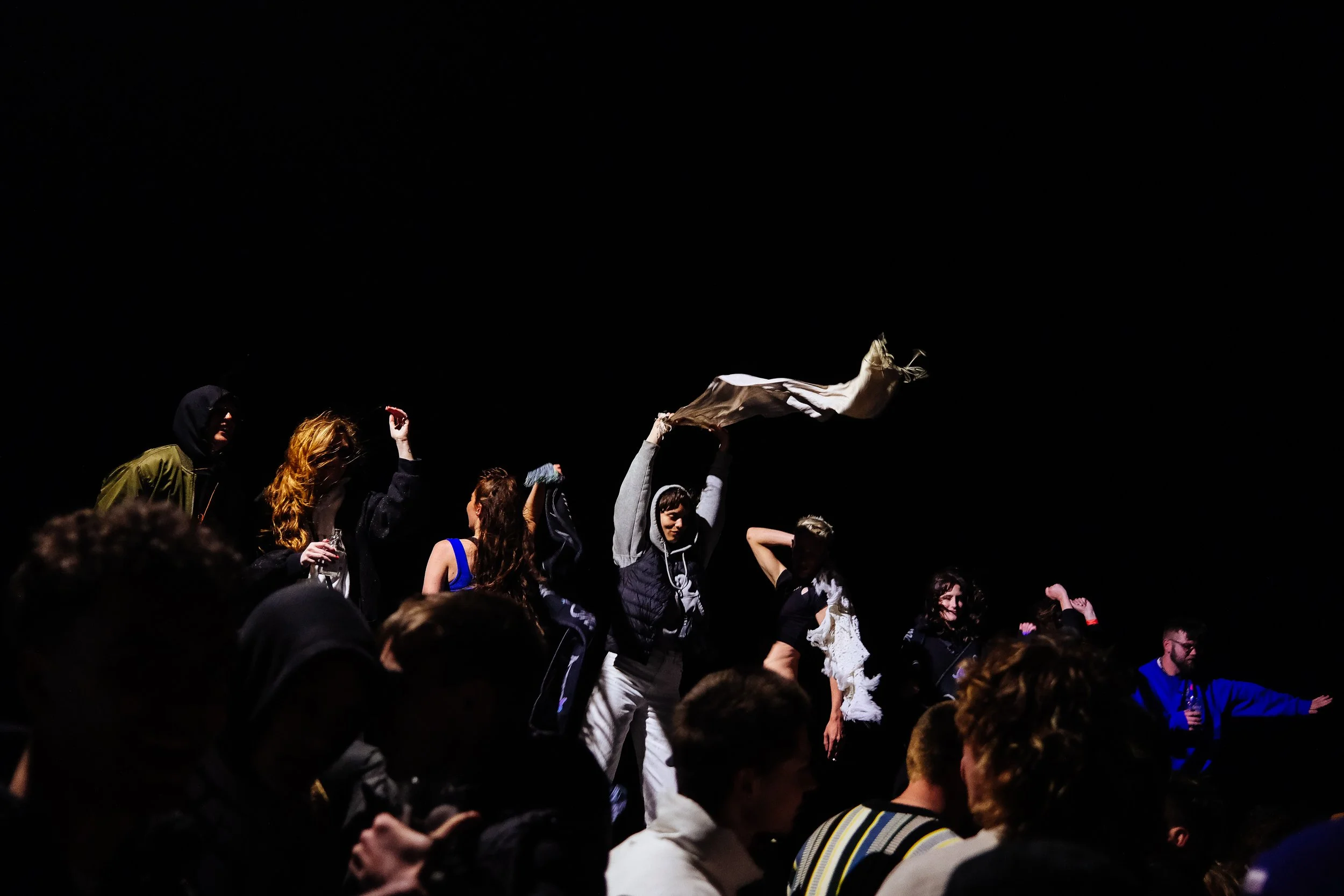 A dark scene with people gathered, some raising their hands, one person waving a cloth, others holding drinks, and smiling, at a lively event or party.