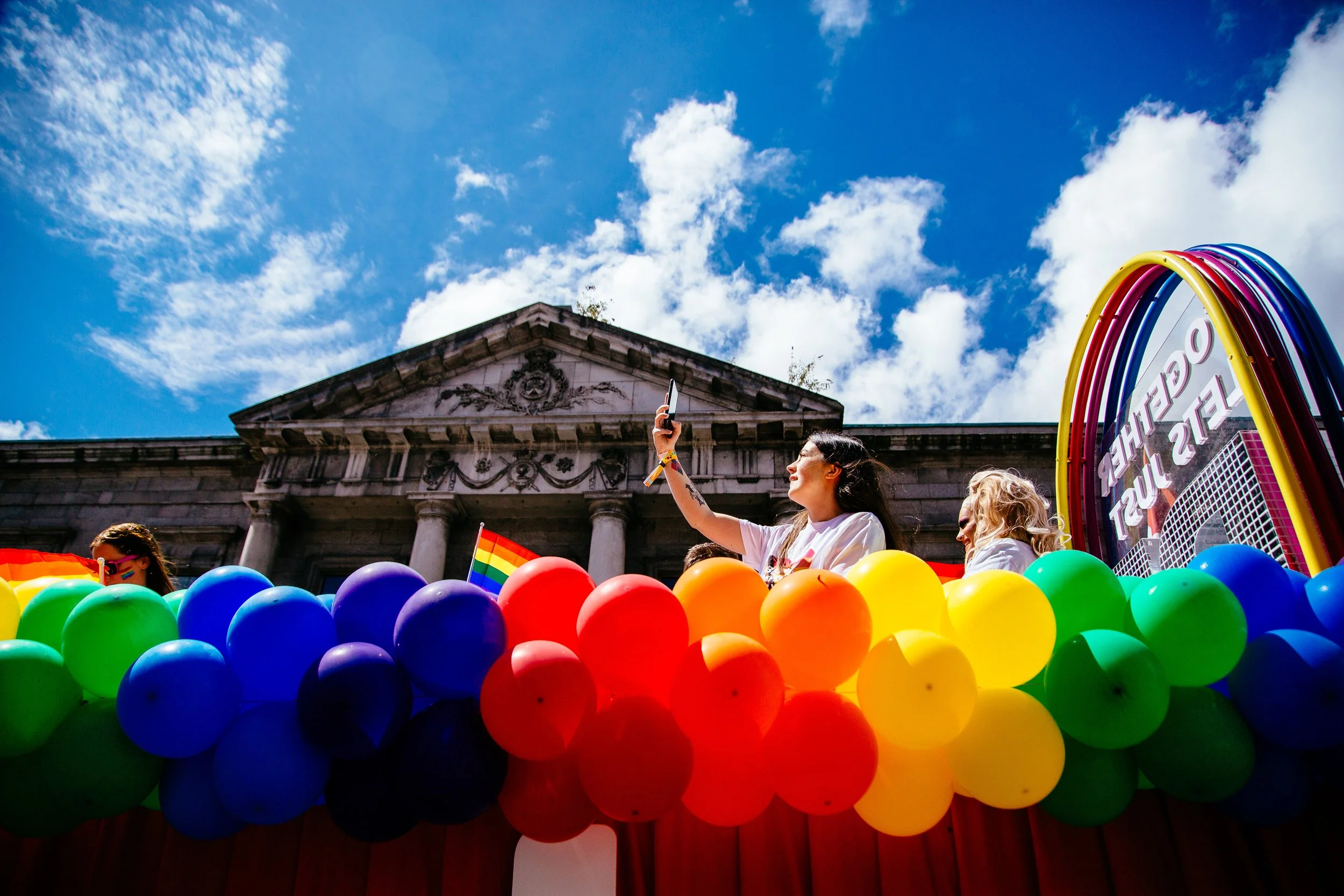 People celebrating at a pride parade with rainbow balloons, holding rainbow flags, in front of a historic building under a blue sky with clouds.