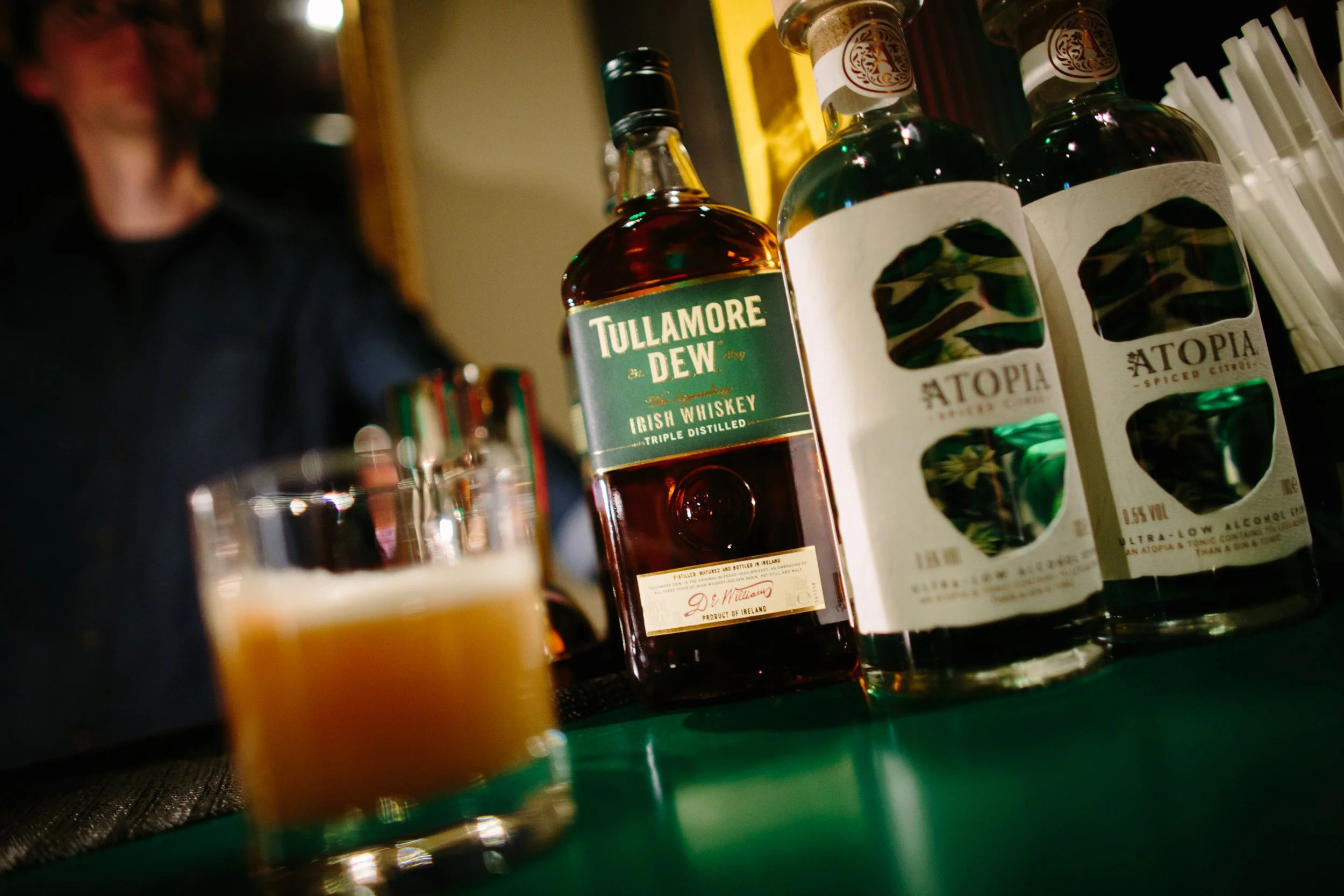 Bottles of alcohol on a table, including Tullamore Dew Irish whiskey and Utopia flavored spirits, with a blurred drink in the foreground and a person on the left side of the background.