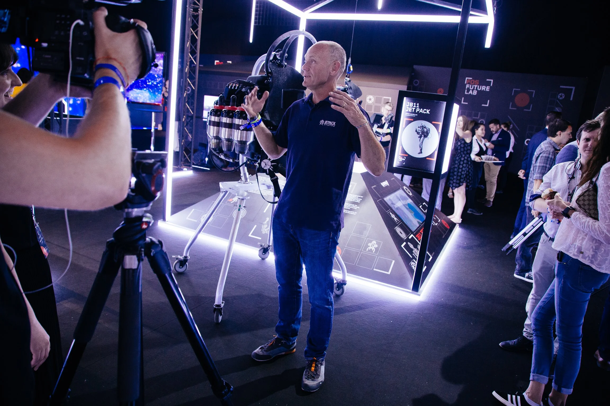 A man in a navy blue shirt with a Jetpack logo is speaking to a photographer at an event. The background has illuminated displays and a sign that reads "FOS Future Lab" and "JB11 Jet Pack." Several people are mingling and examining exhibits.