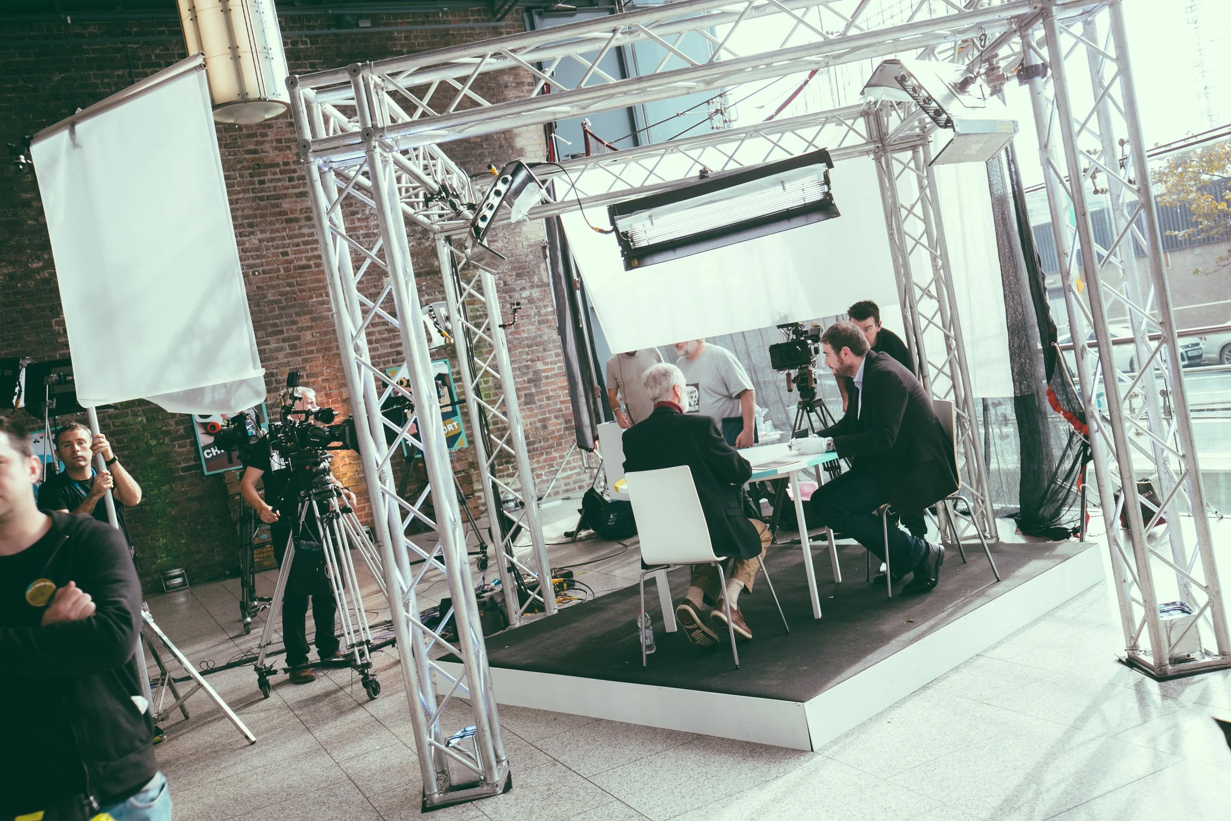 A television interview setup inside a building with brick walls. There are two men sitting at a table being filmed or interviewed, with professional lighting and camera equipment. Crew members are visible, one holding a reflectors, and others operati