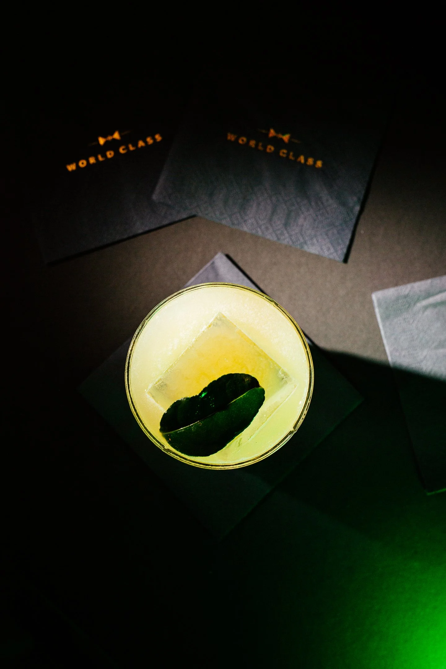 A glass of yellow-colored drink garnished with a slice of lime and mint leaves on top, with a dark background and black cards with gold text that read 'World Class'.