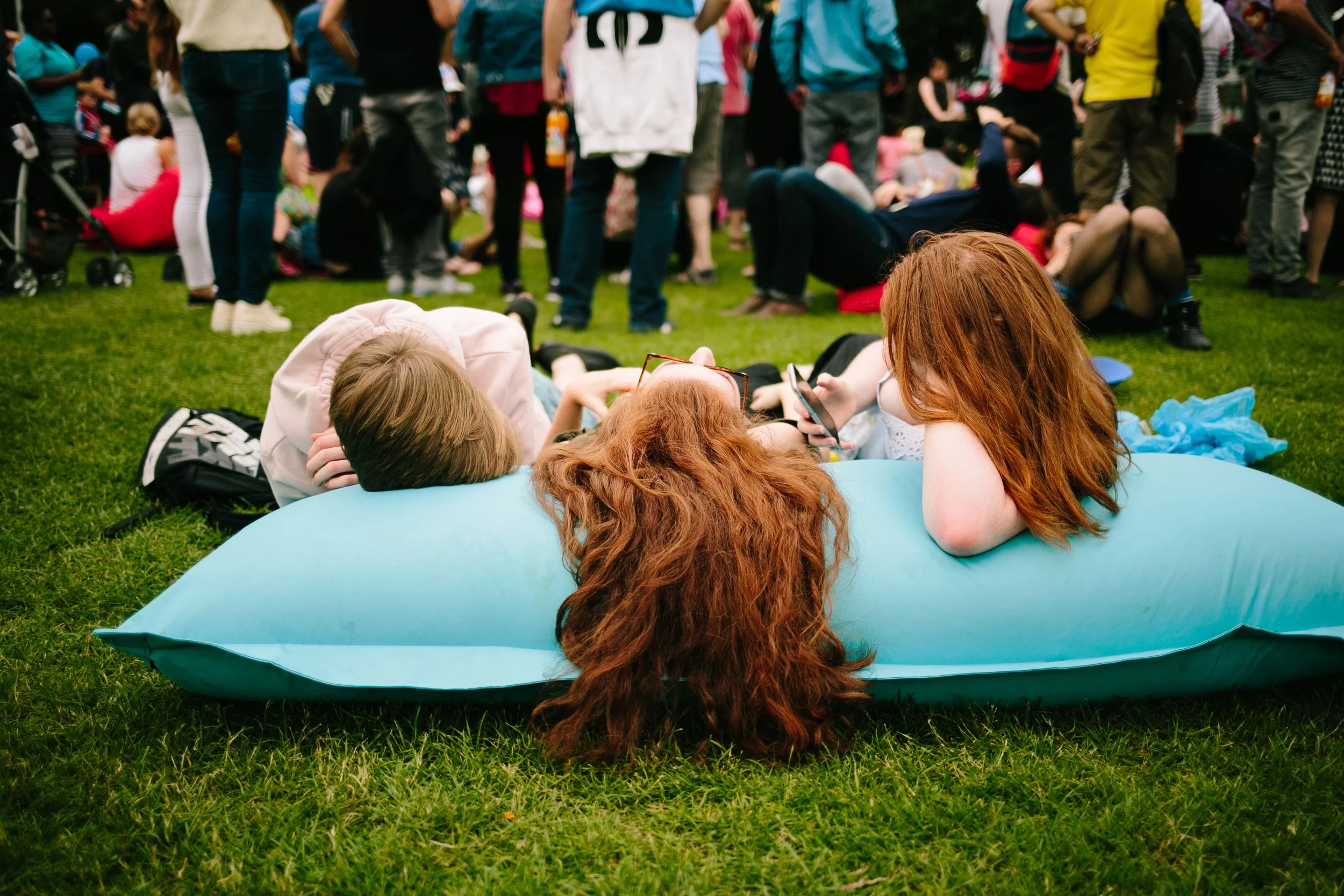 People gathered outdoors on a grassy field, sitting or lying down, with some standing in the background. Two women with red hair are lying on a large blue inflatable object, looking at a smartphone together. The crowd appears to be at a social or rec