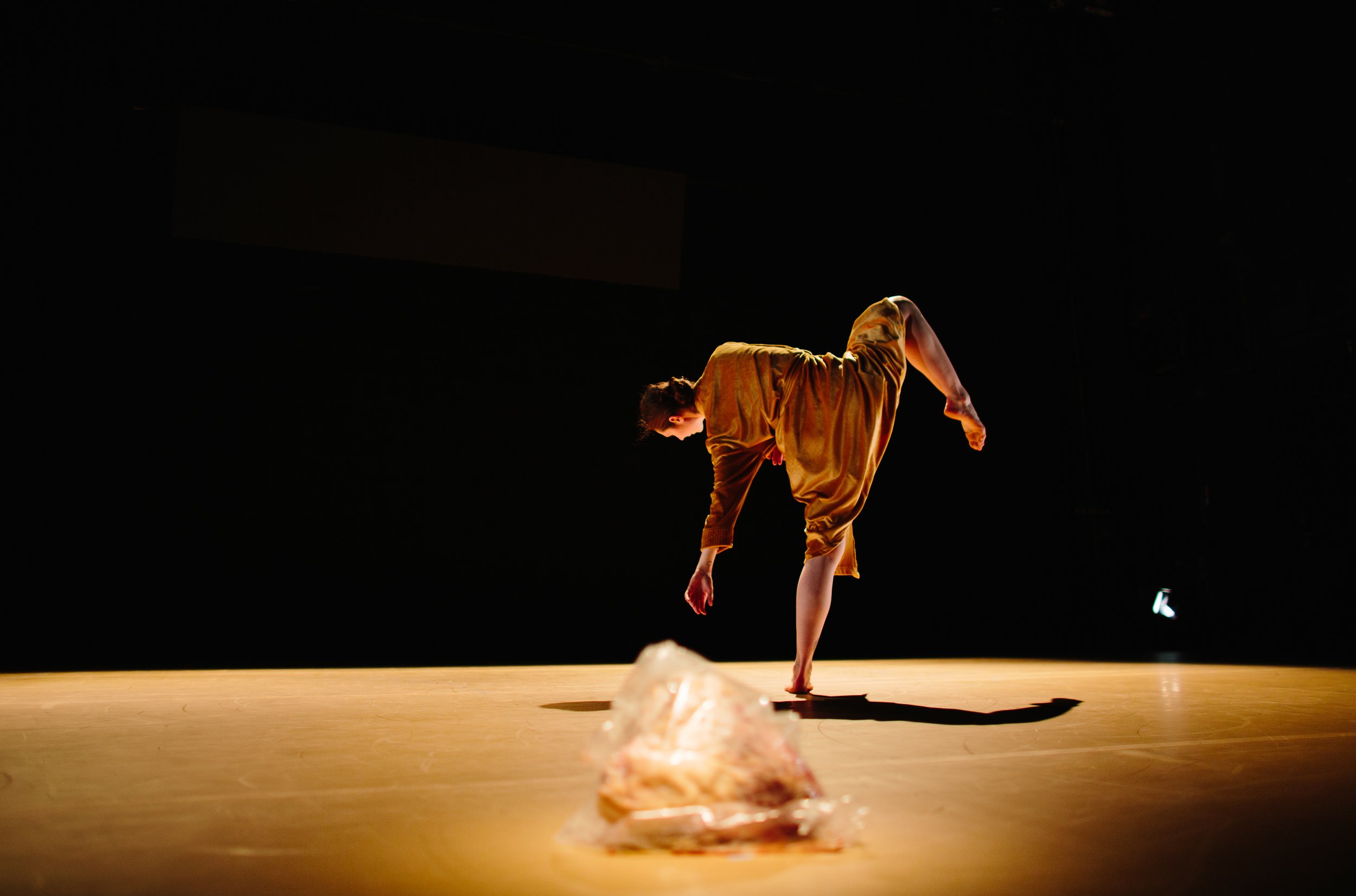 A person performing a dance move on stage, bending forward with one leg raised and their hand touching the floor in a spotlighted setting.